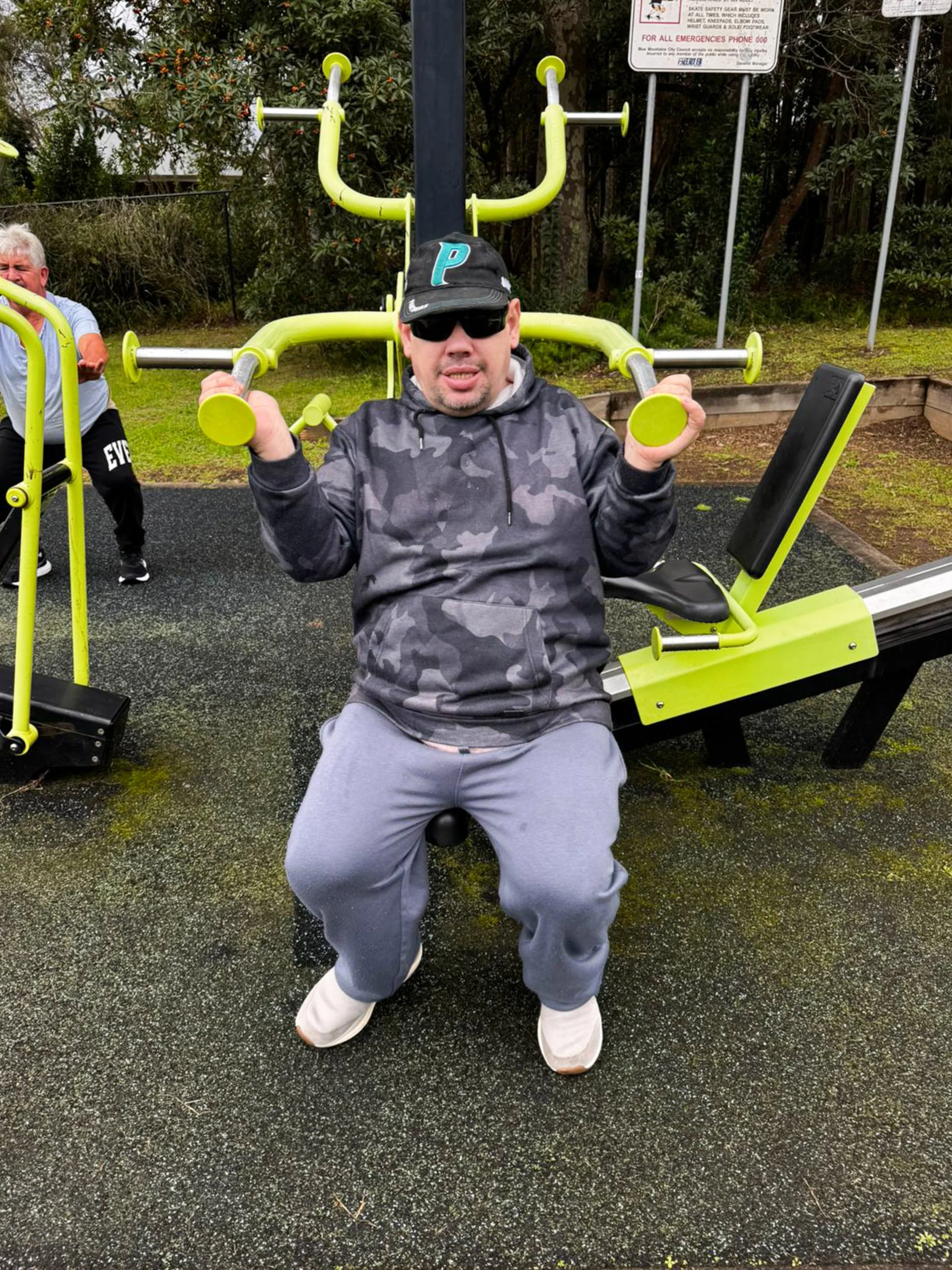 Man sitting on a fitness ball in an outdoor gym, lifting light green exercise dumbbells, wearing a black camouflage hoodie, gray sweatpants, a black cap with a blue background letter P, and sunglasses. There is outdoor fitness equipment and another p
