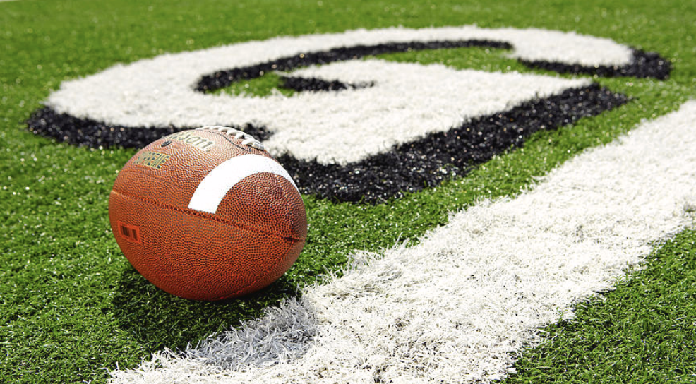 A Wilson football on a green turf field near a painted logo with black and white elements.