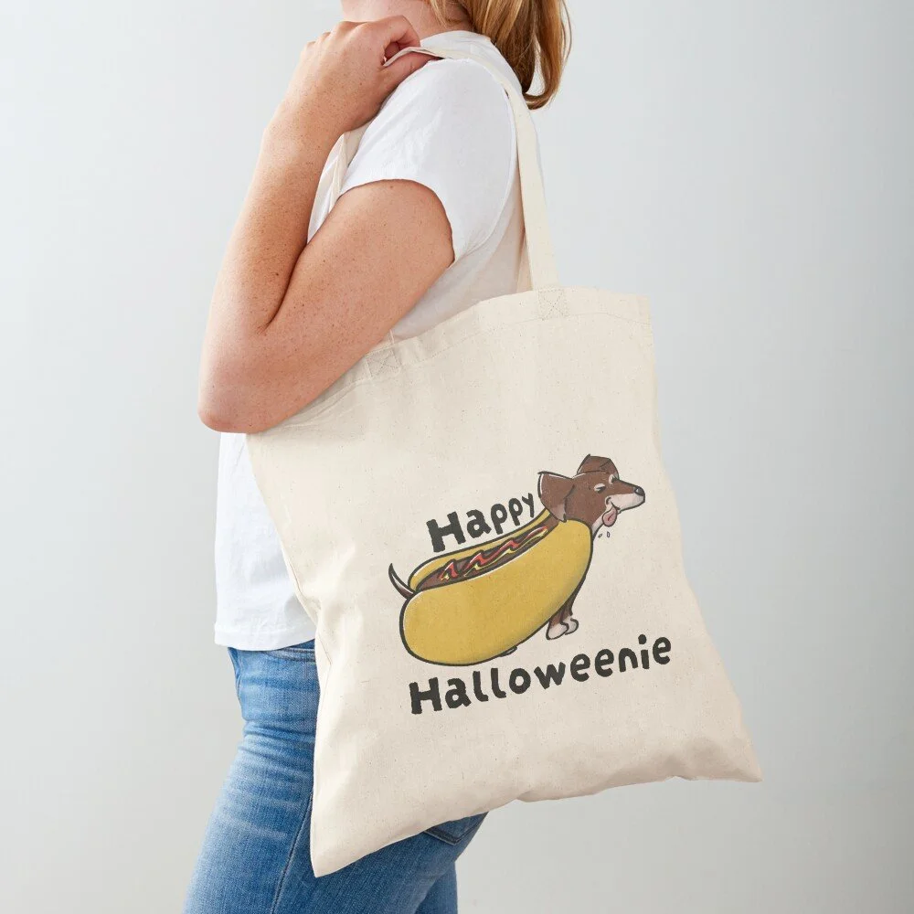 Person wearing a white t-shirt and blue jeans carrying a tote bag with a Halloween-themed cartoon dog inside a hot dog bun with the words "Happy Halloweenie".