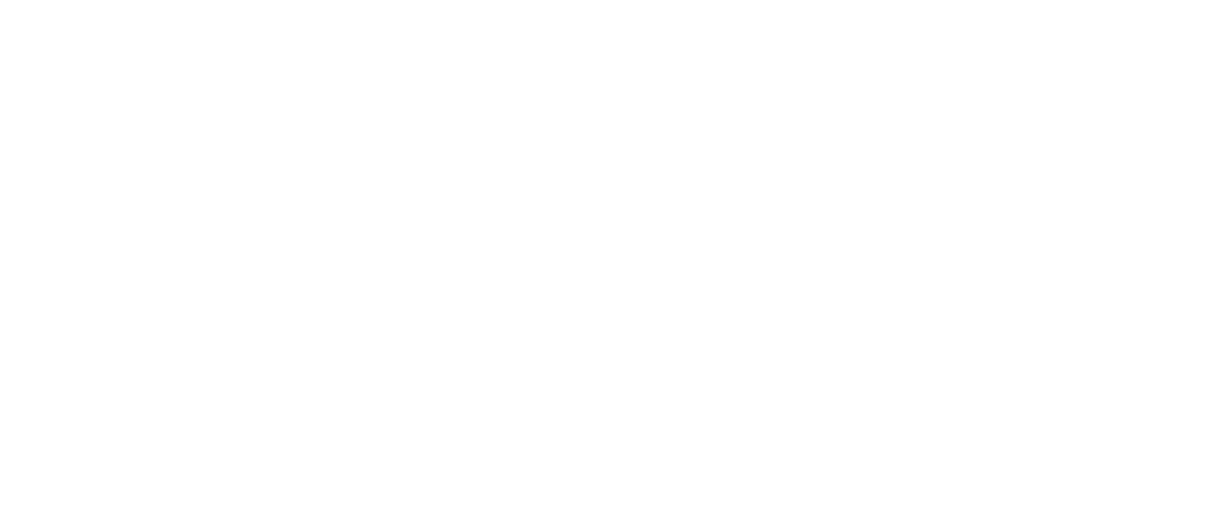 A person wearing a white shirt and jeans, riding a motorcycle on a bridge over a canal with lush green trees and buildings in the background.