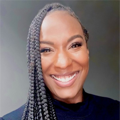 Close-up of a smiling woman with braided hair and dark clothing, against a neutral background.