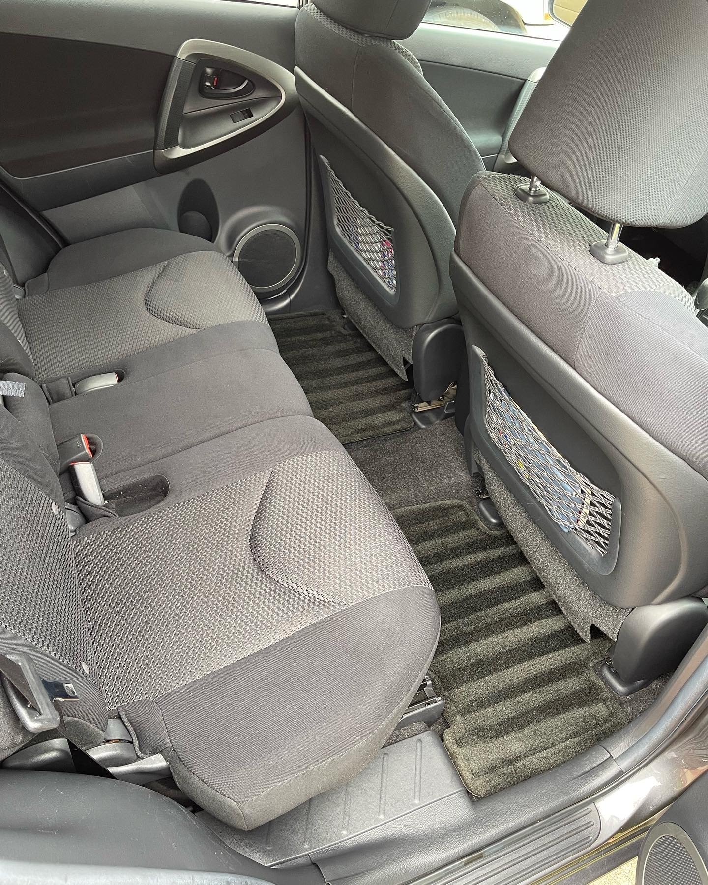 Interior of a vehicle showing the back seat area with gray fabric seats, floor mat, and back of front seats with mesh pockets.