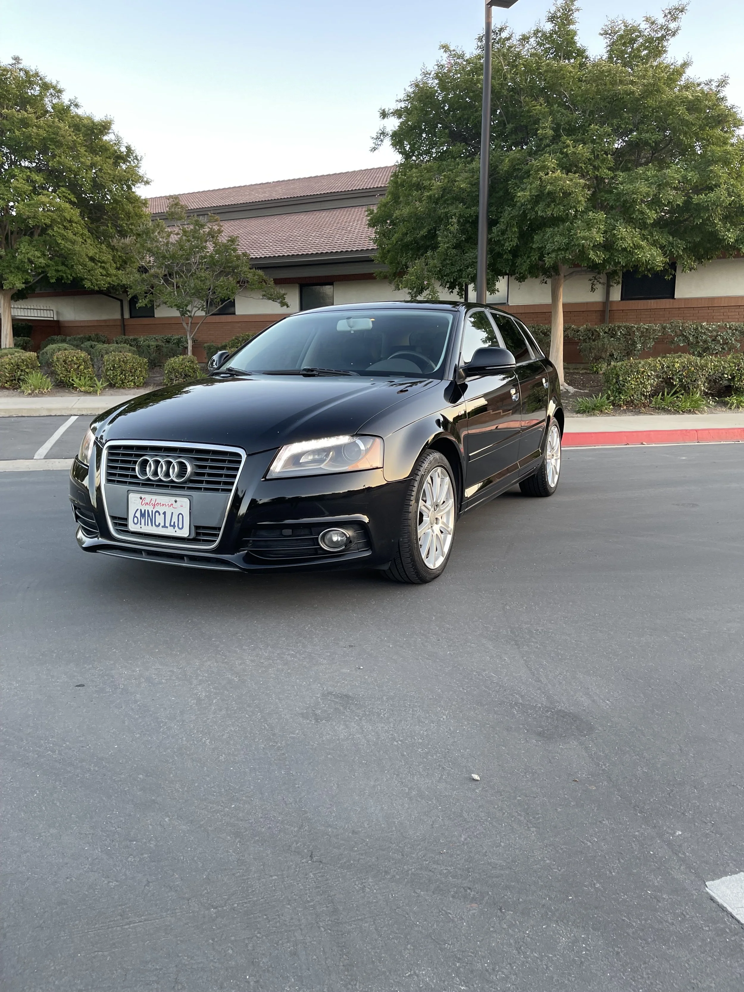 Black Audi sedan parked in a parking lot with trees and a building in the background.