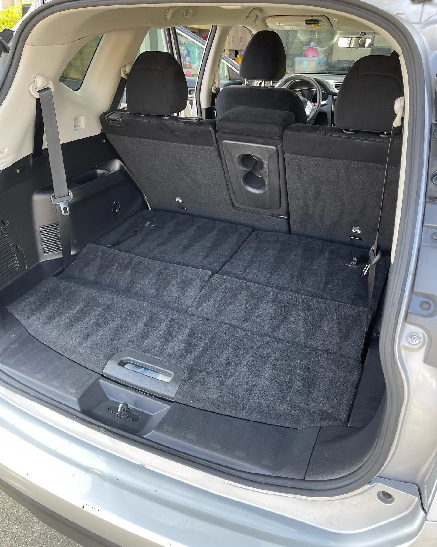 The photo shows the interior of an SUV with the rear seats folded down, creating a flat cargo area covered with black carpeting. The front seats are visible through the open rear hatch, and there are some objects and decorations visible outside the vehicle.