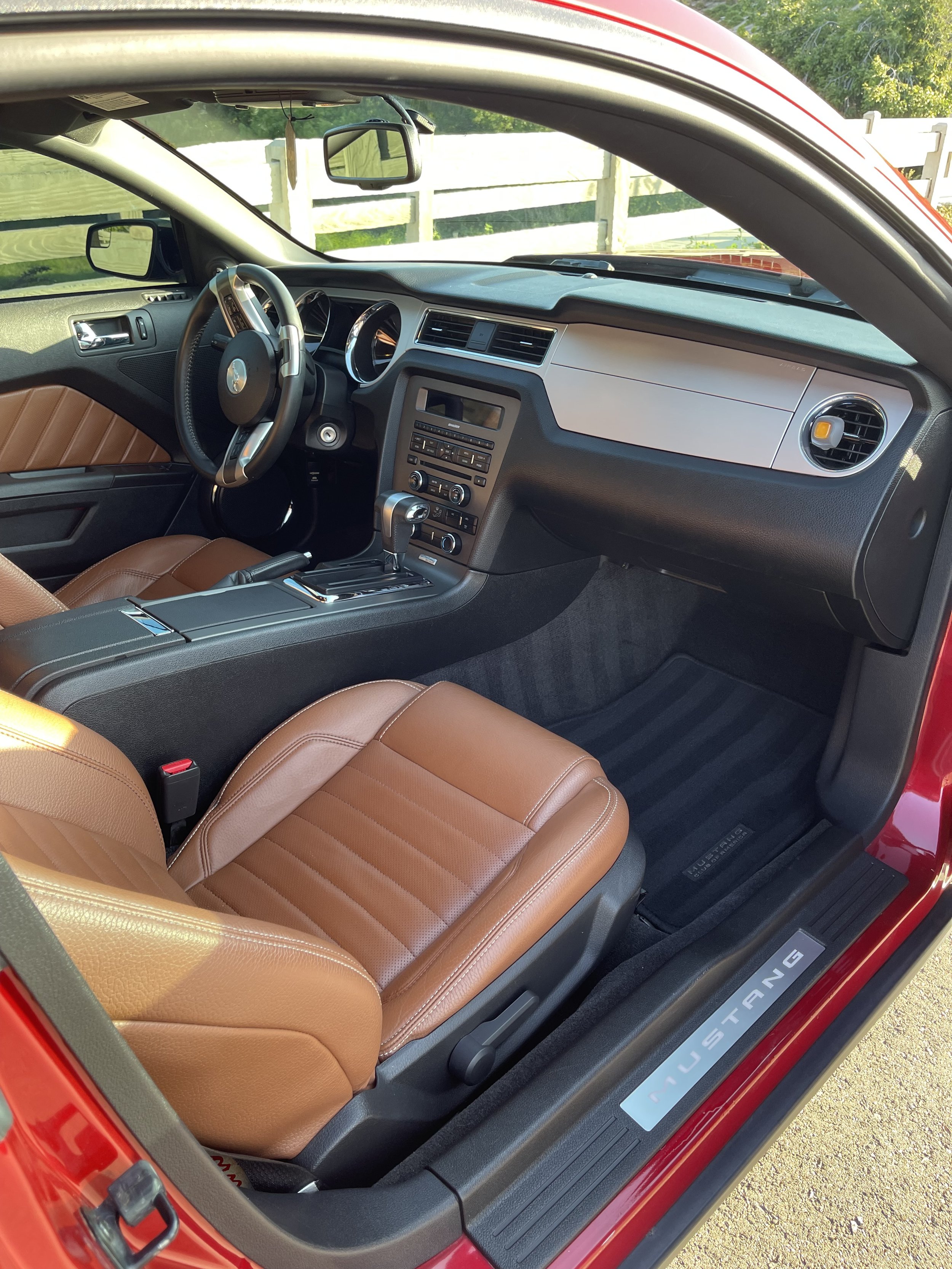 Interior of a red Ford Mustang with brown leather seats, dashboard, and center console, parked outdoors near a wooden fence.