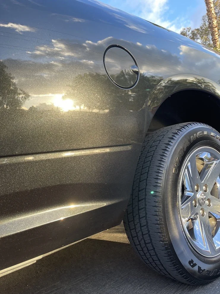 Close-up of the side of a black pickup truck showing a gold metallic paint, the fuel cap, and a large tire with a chrome rim parked outdoors during sunset or sunrise with trees and a partly cloudy sky reflected on the surface.