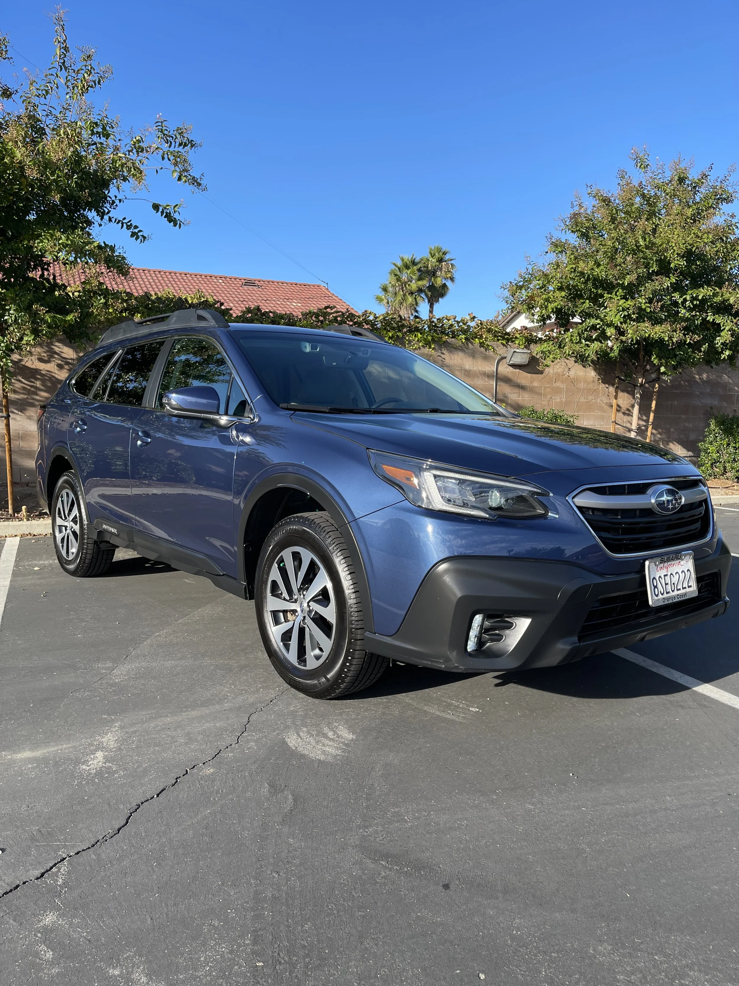 Blue Subaru Outback parked in a parking lot with trees and a brick wall in the background.