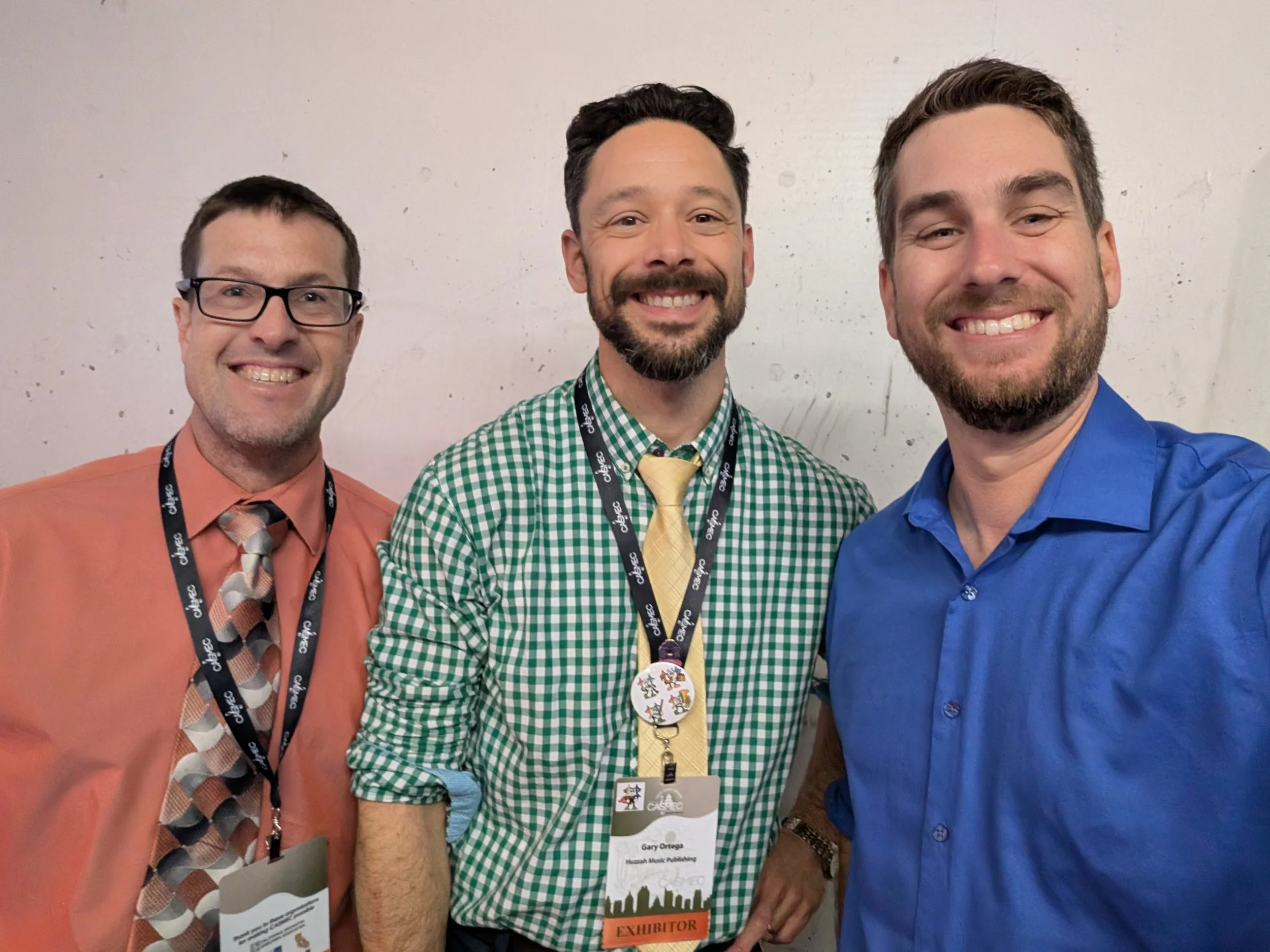 Three men dressed in colorful shirts smiling at the camera, wearing conference badges, standing in front of a plain wall. Keith Hunter, Gary Ortega, and Shawn McGinn