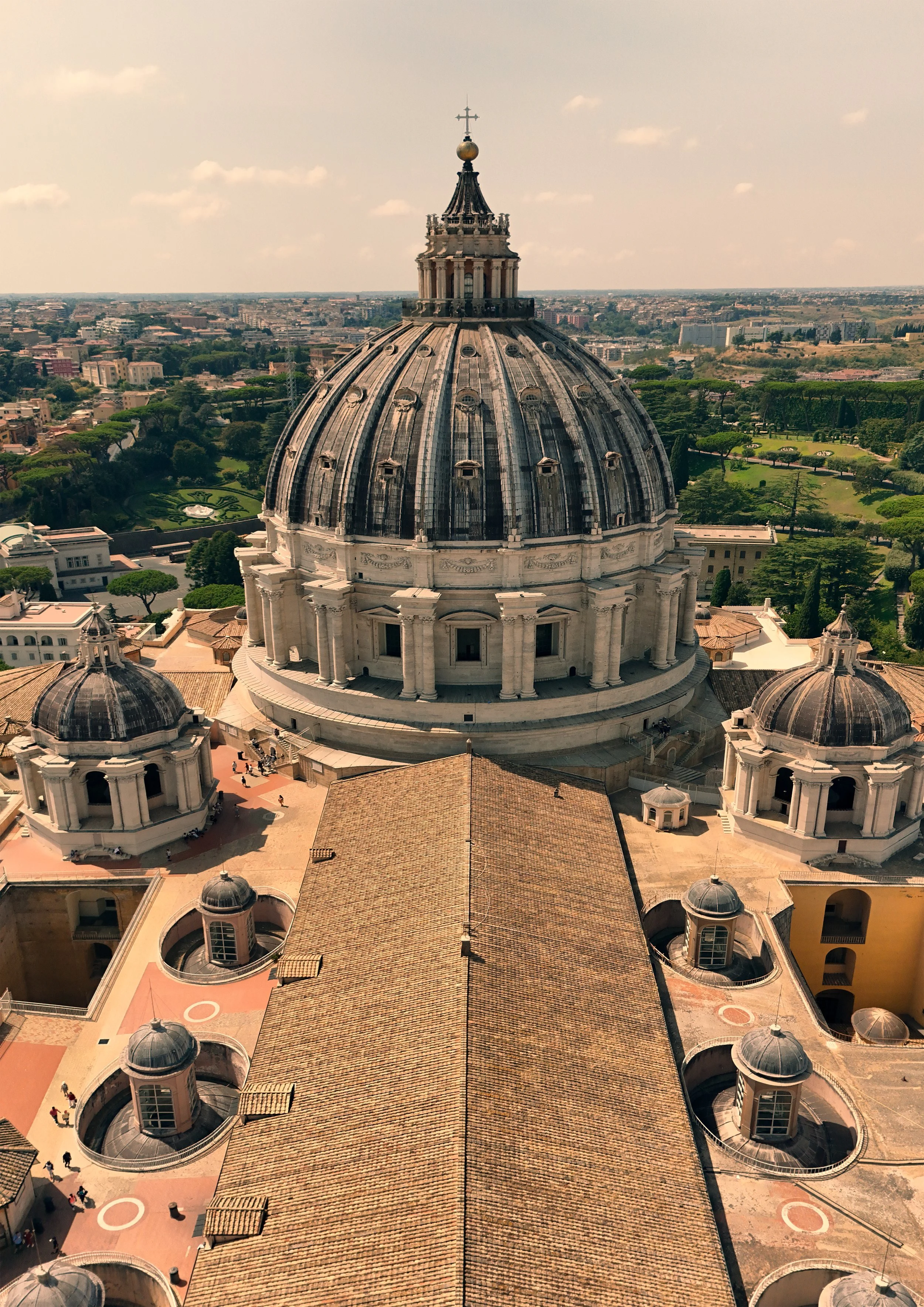 Aerial view of the large dome of St. Peter's Basilica in Vatican City, with smaller surrounding domes and a rooftop, and a cityscape in the background under a partly cloudy sky.