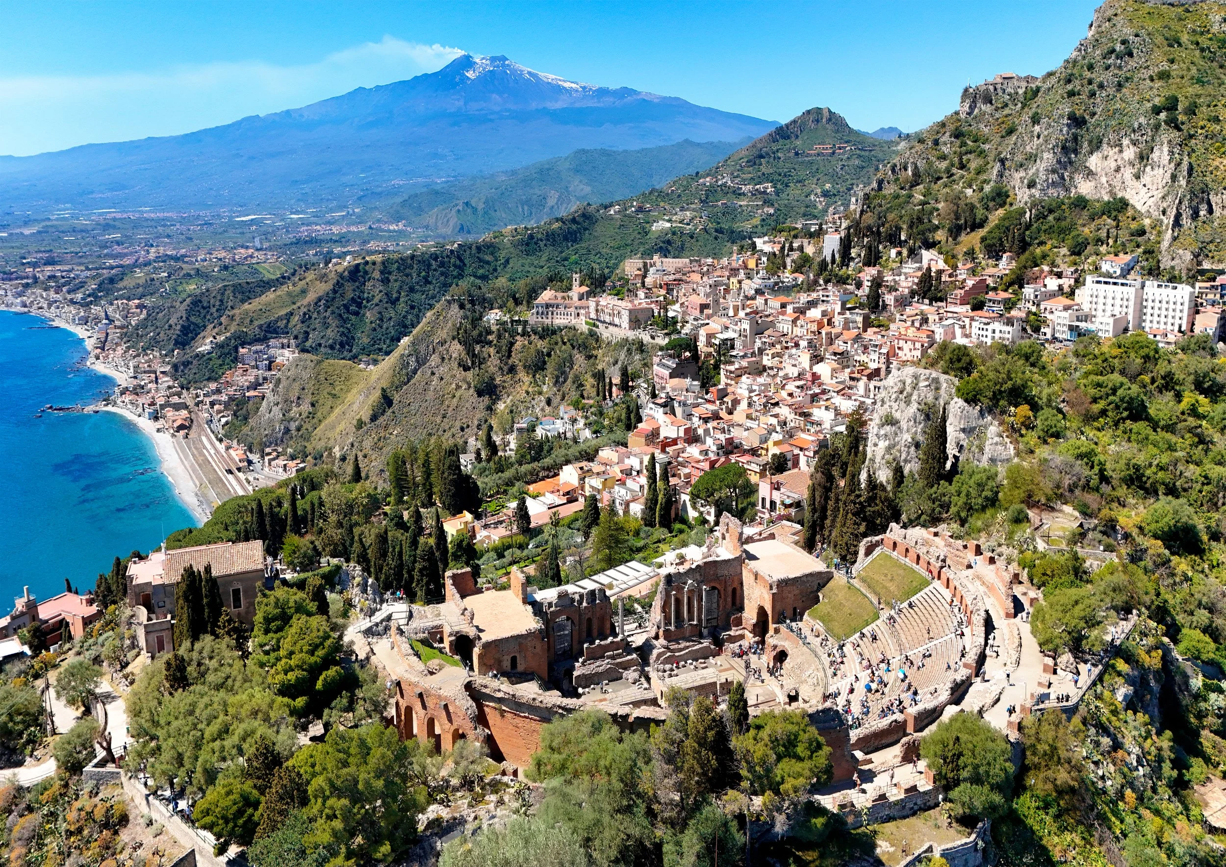 A coastal hillside with ancient ruins, a Mediterranean town with colorful buildings, the ocean with a beach, and a snow-capped mountain in the background.