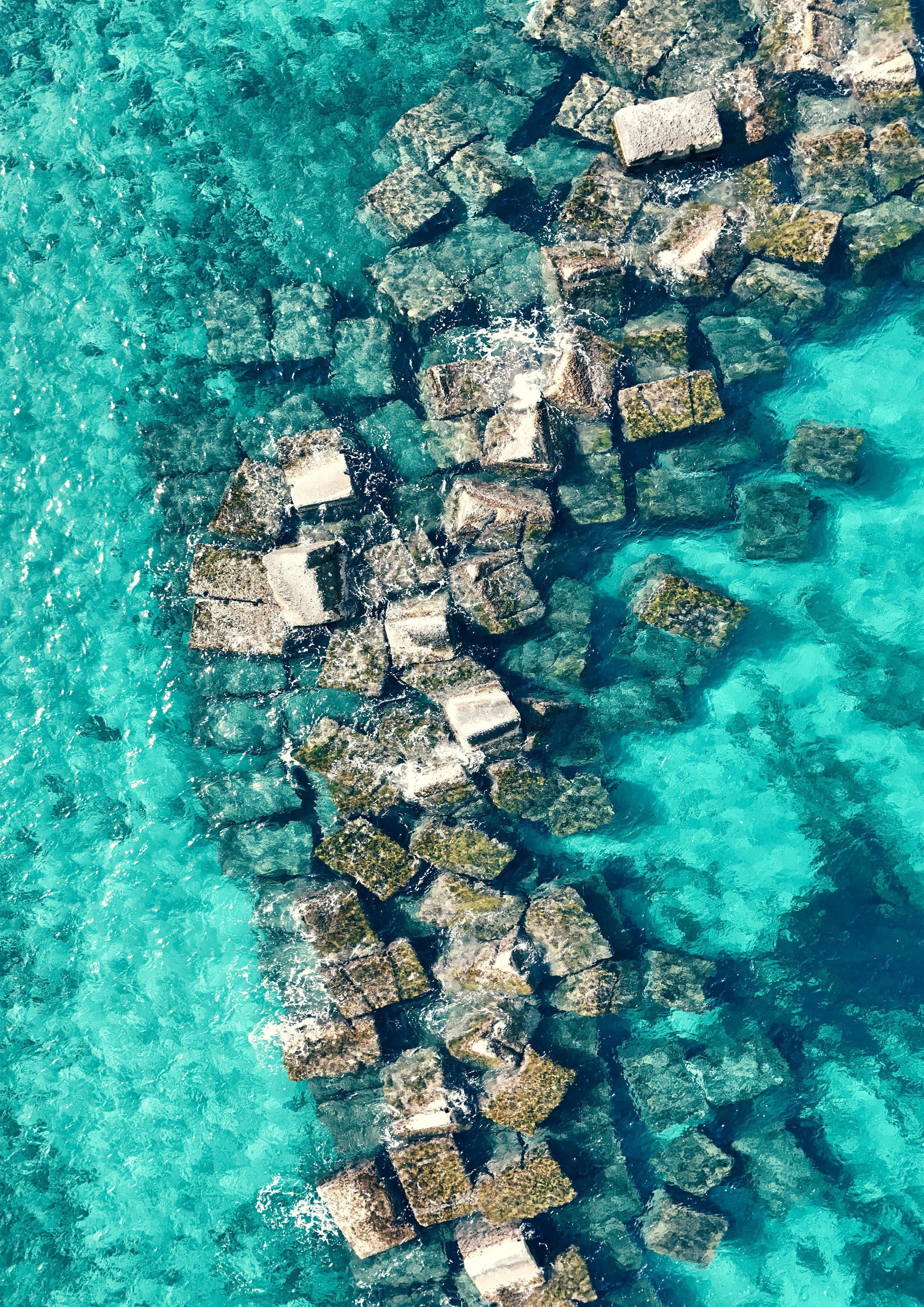 Aerial view of a line of large rocks or concrete blocks extending into the ocean, creating a breakwater or jetty, with clear turquoise water surrounding them.