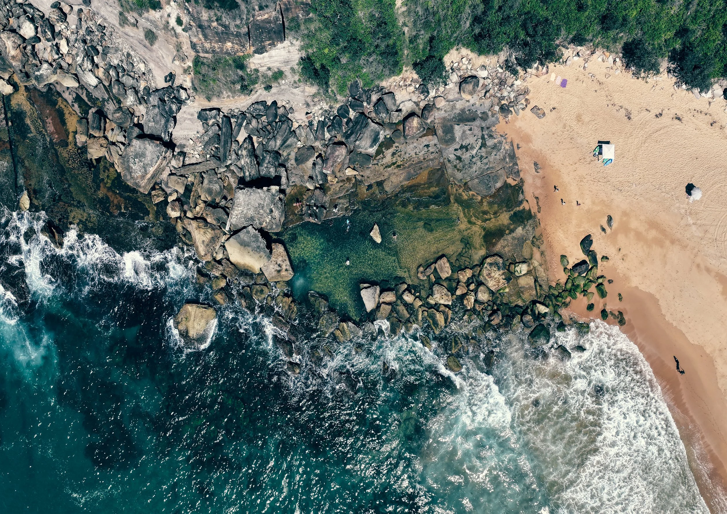 An aerial view of a beach with rocky shoreline, sandy area with umbrellas, and ocean waves crashing against rocks.
