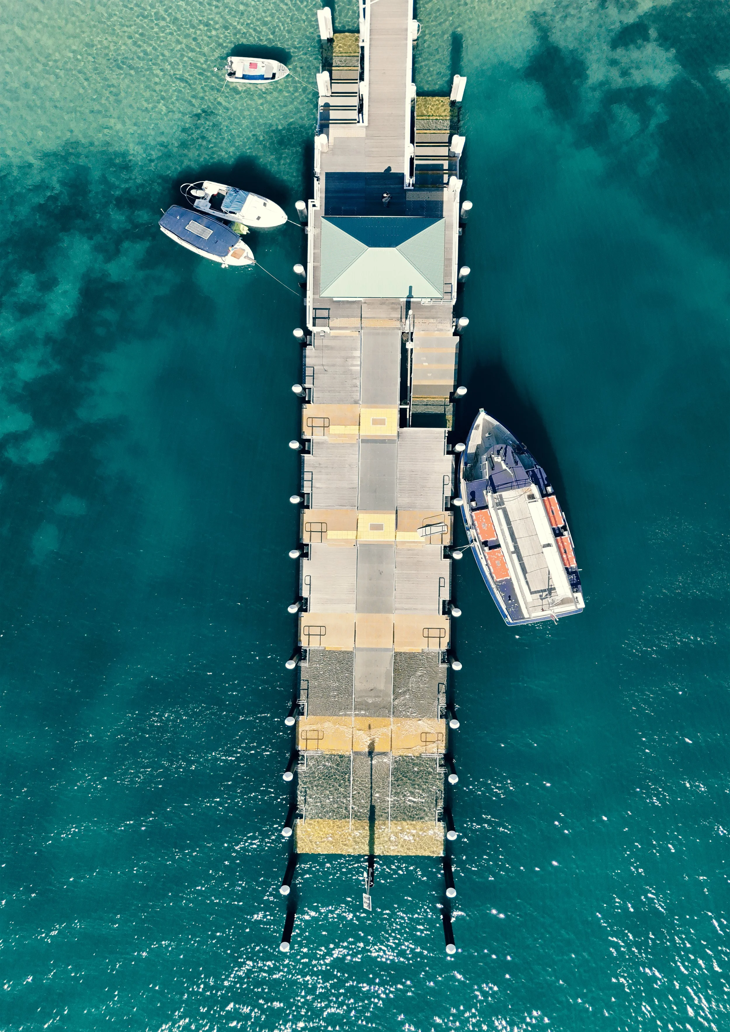 Aerial view of a boat dock extending into the water with three boats moored alongside, on clear turquoise water.