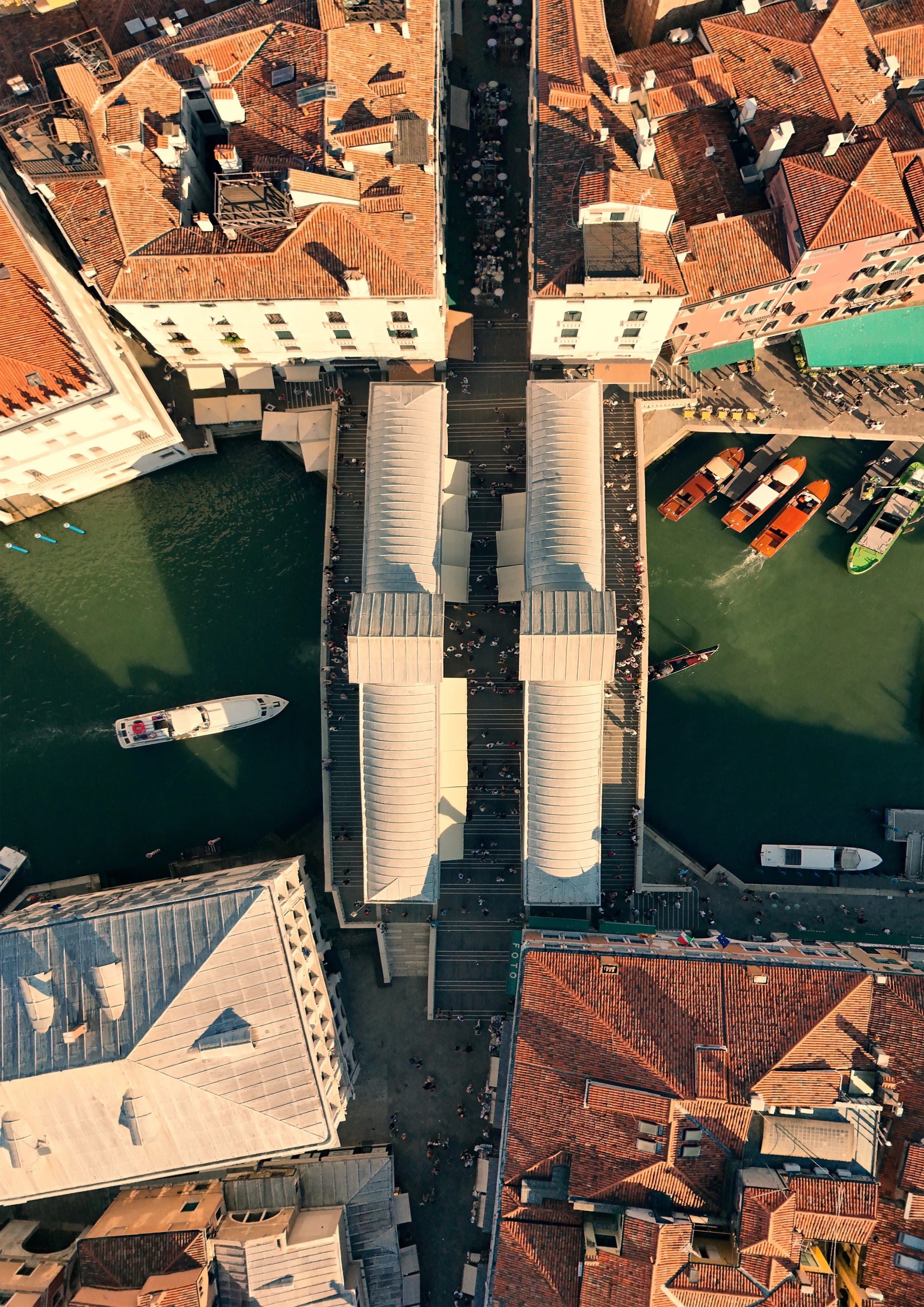 An aerial view of a city canal with boats, surrounding buildings with red and gray roofs, and a crowded pedestrian bridge over the water.