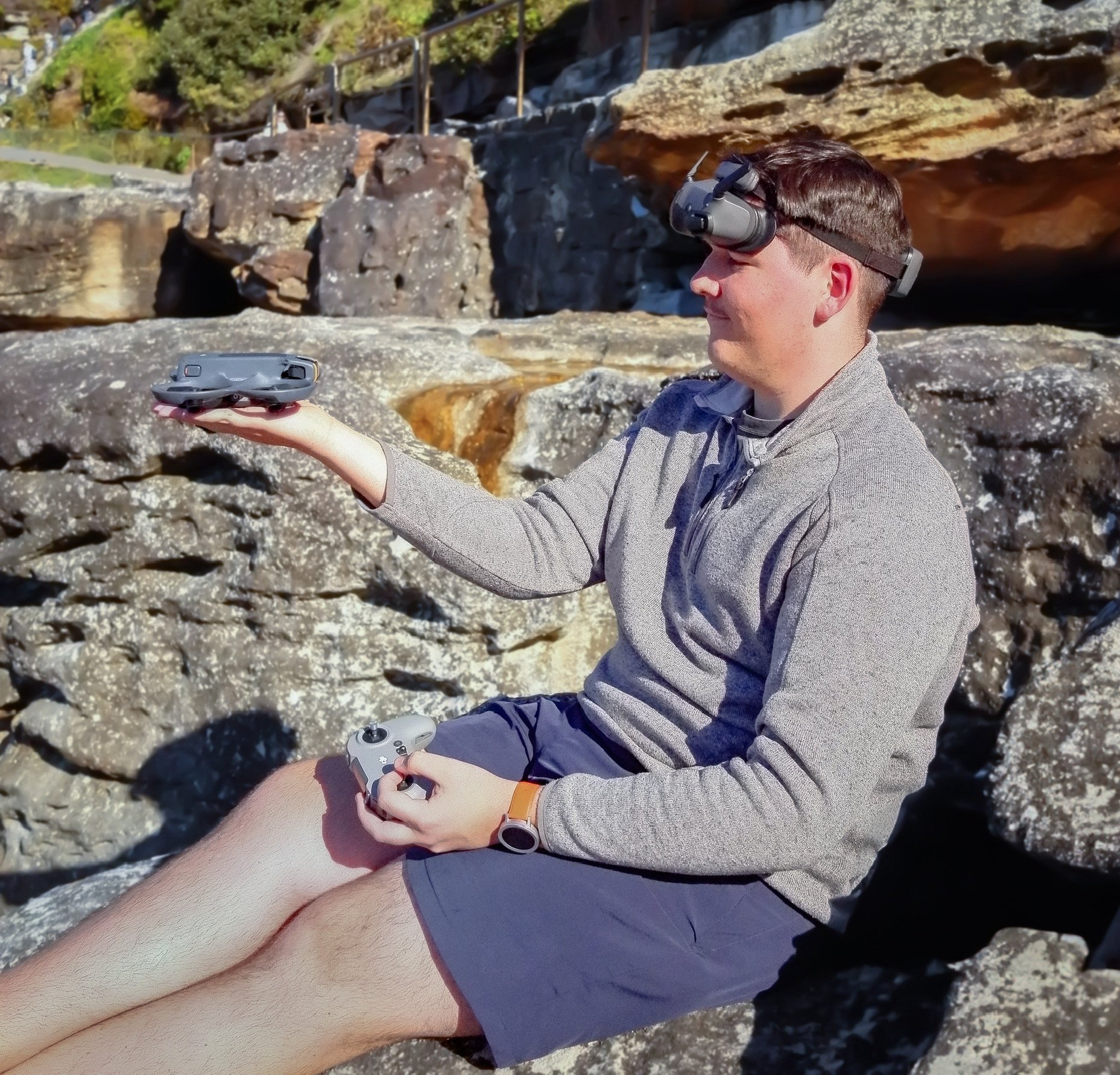 A man sitting on rocks outdoors, holding a drone and a remote control, wearing a drone headset, gray jacket, and shorts.