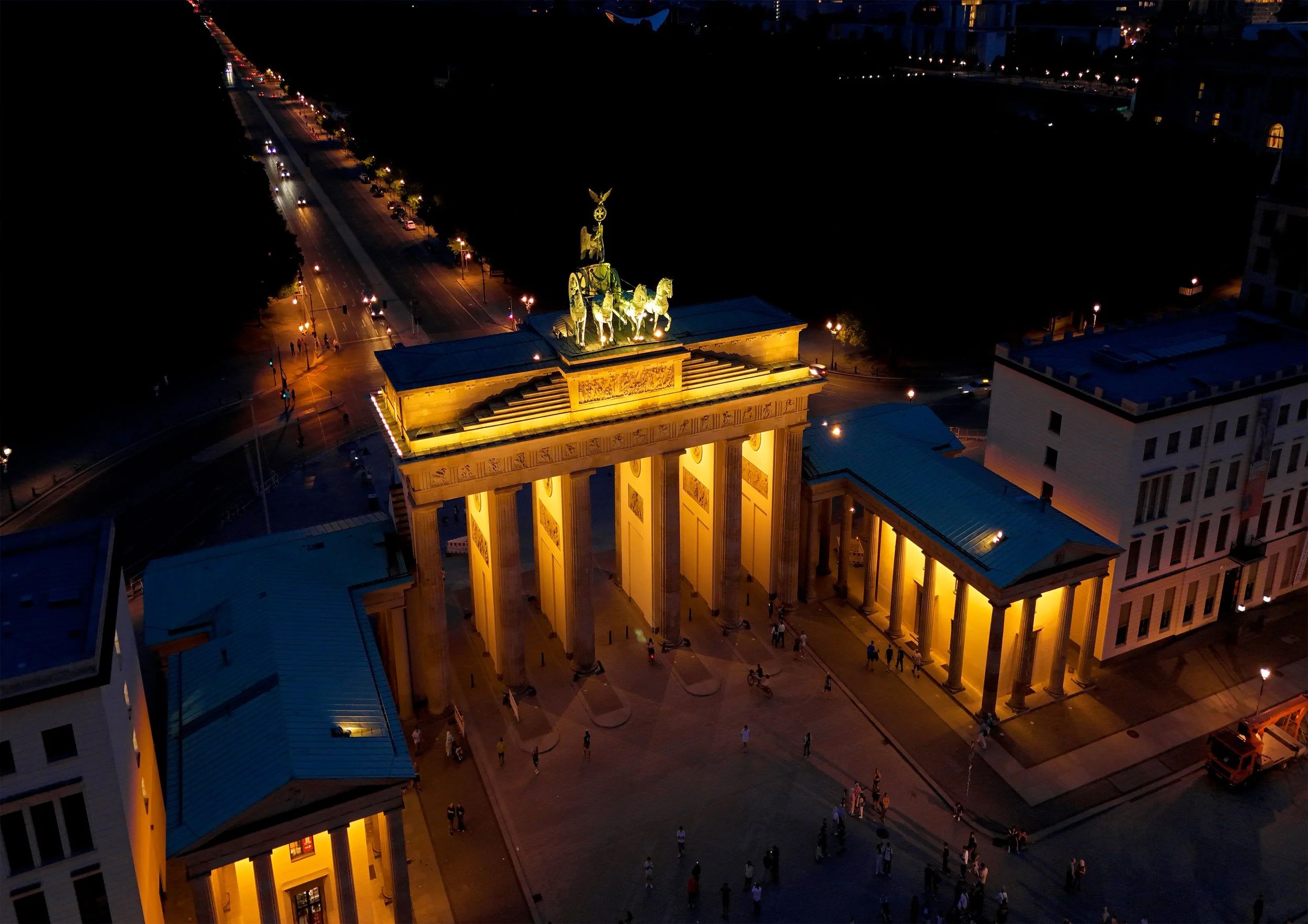 Night aerial view of the Brandenburg Gate illuminated, with people walking around and city streets visible in the background.