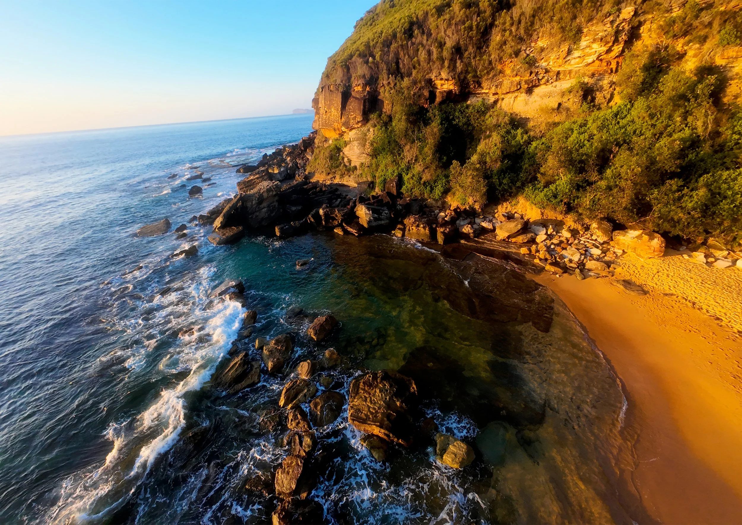 Cliffside coastline with rocky shore, waves, sandy beach, and green vegetation, illuminated by golden sunlight at sunset.