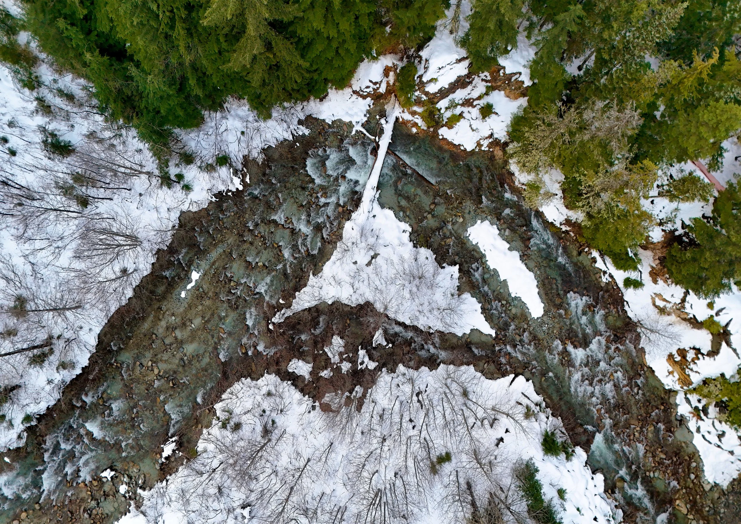 An aerial view of a winding river through a snow-covered forest with evergreen and deciduous trees.