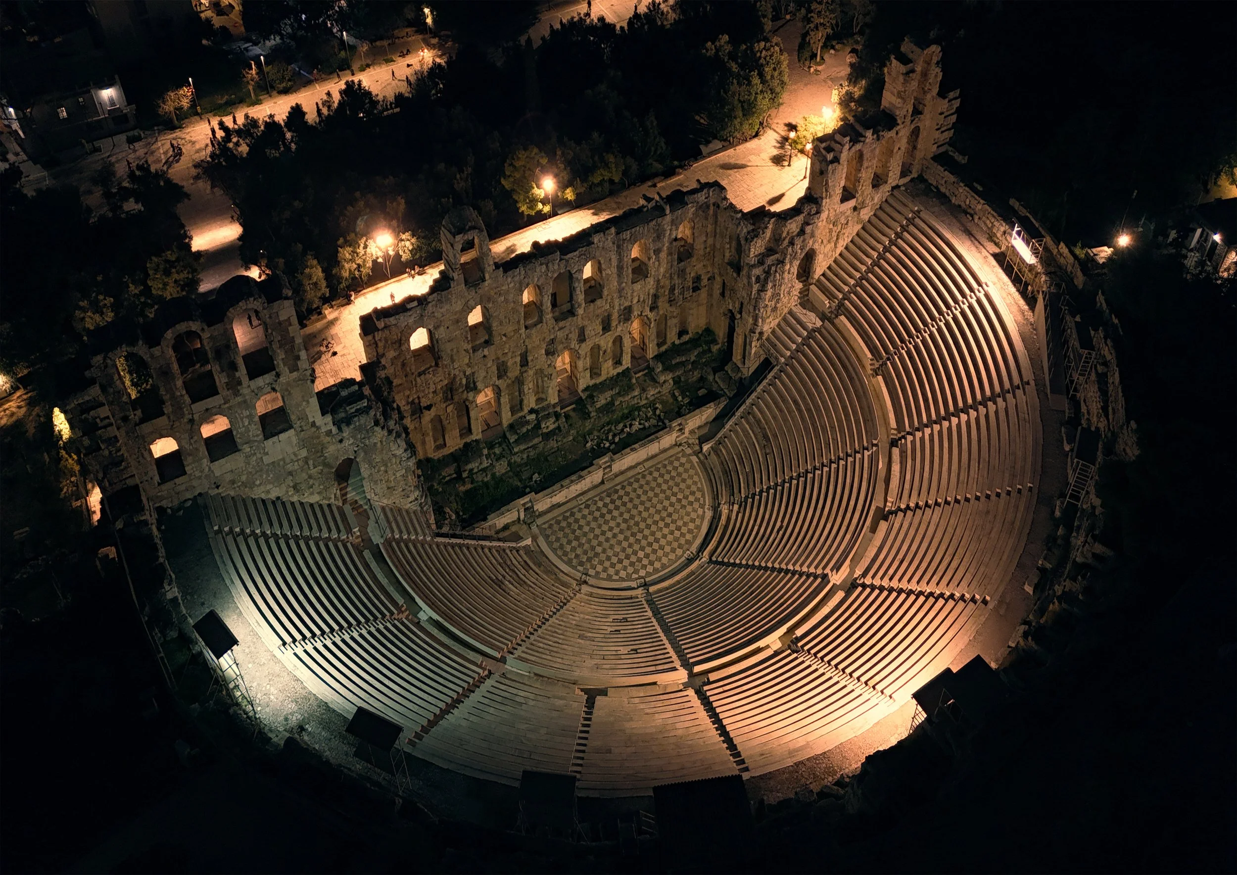 An aerial night view of an ancient outdoor theater with curved rows of seats, a checkered stage, and a partially ruined stone wall behind it, illuminated by surrounding lights.