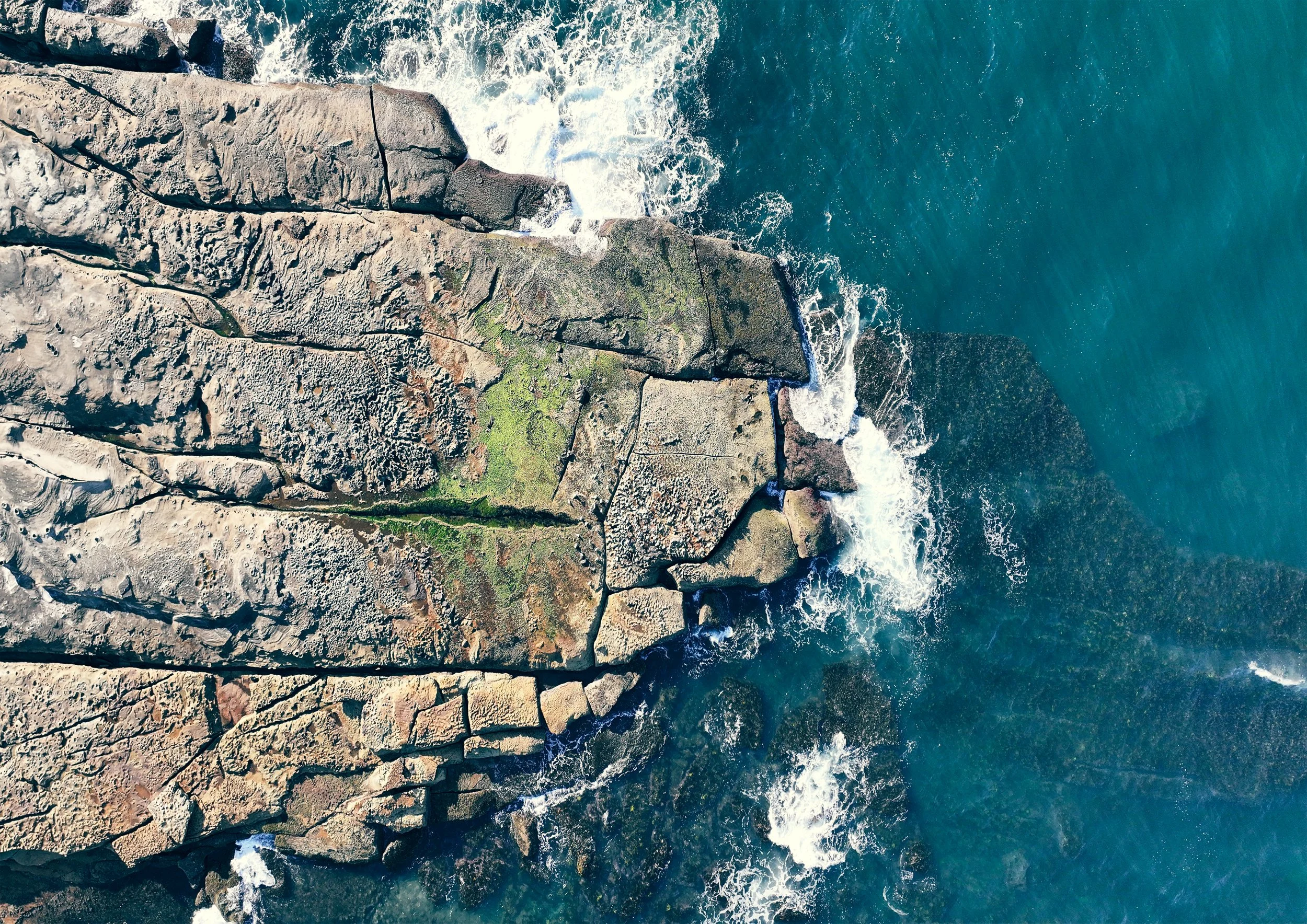 Aerial view of a rocky shoreline with green moss, waves crashing against rocks, and the ocean extending to the right.