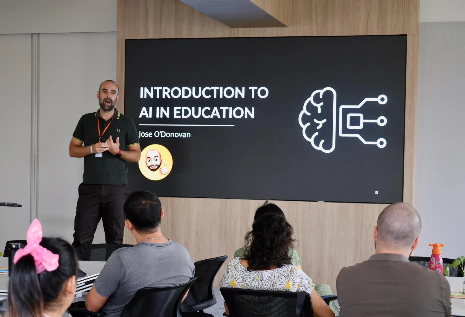 Man giving a presentation on AI in education to a seated audience in a conference room with a large screen behind him showing a brain and circuit icon, and the title 'Introduction to AI in Education' by Jose O'Donovan.