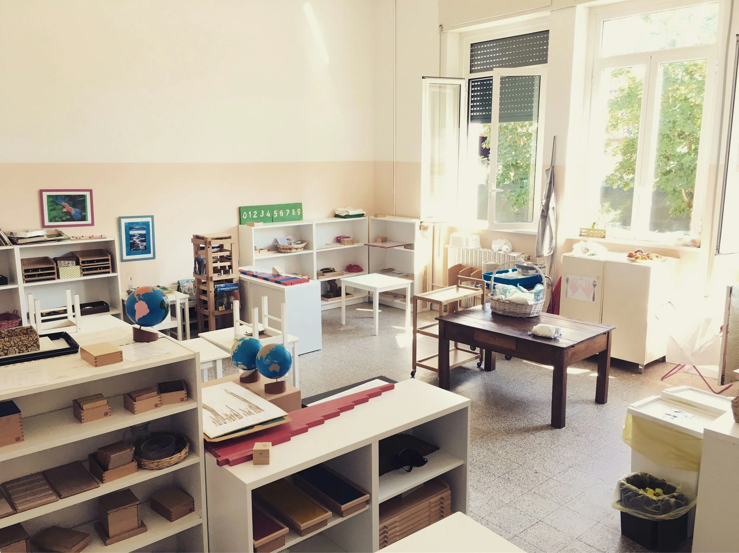 A bright classroom or playroom with shelves filled with educational toys, including globes, blocks, and wooden puzzles. Large windows let in natural light, revealing green trees outside.