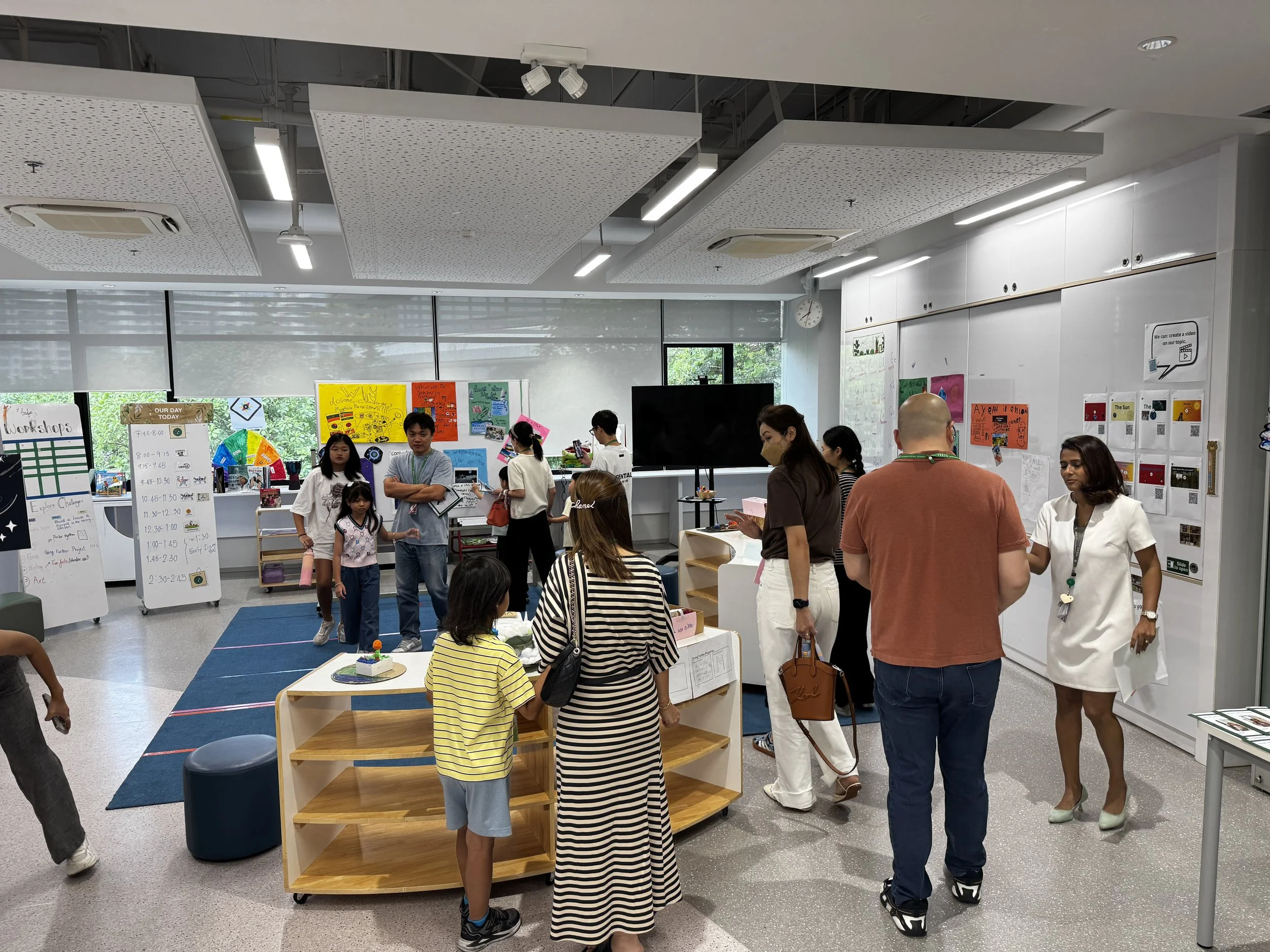 A classroom or event space with people, including children and adults, engaging in activities. Posters and displays are on the walls and tables, with some people looking at materials and others talking. The room has white walls, large windows with shades, and a ceiling with lights and air conditioning units.