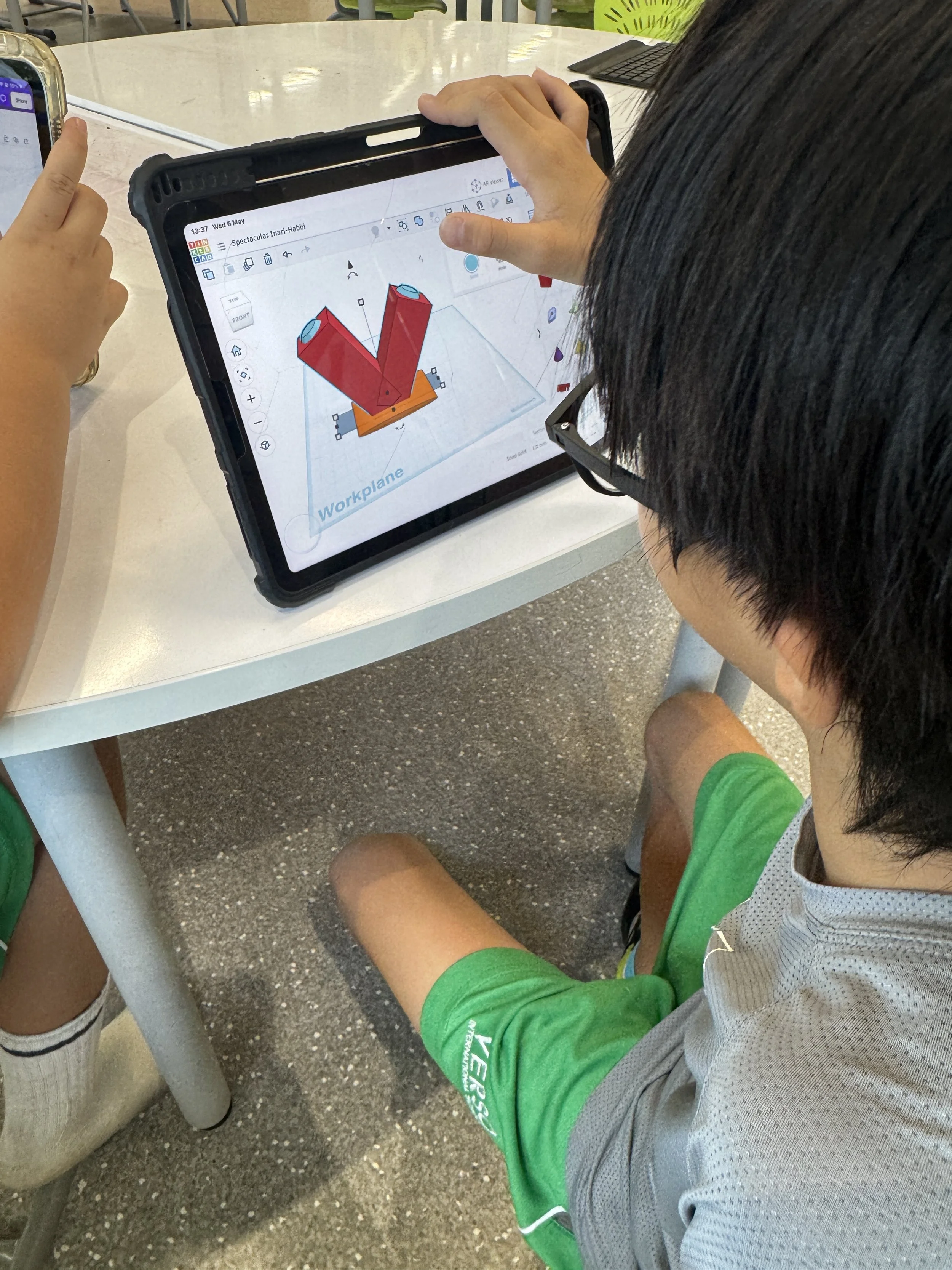 A person wearing a gray and green athletic shirt and shorts is sitting at a white table, looking at a tablet with a 3D design of red and orange objects on the screen, in an outdoor setting.