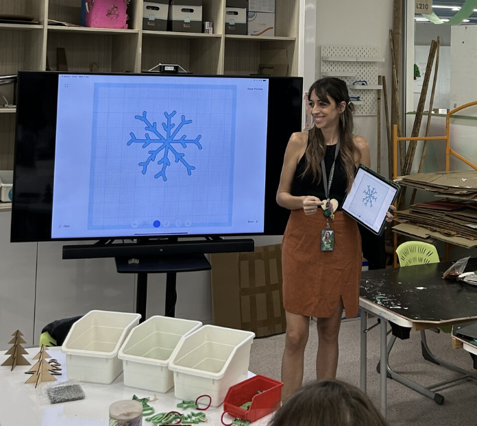 Woman standing in a classroom holding a tablet displaying a snowflake graphic, with a large screen behind her showing the same snowflake image. There are craft materials and small paper trees on the table in front of her.