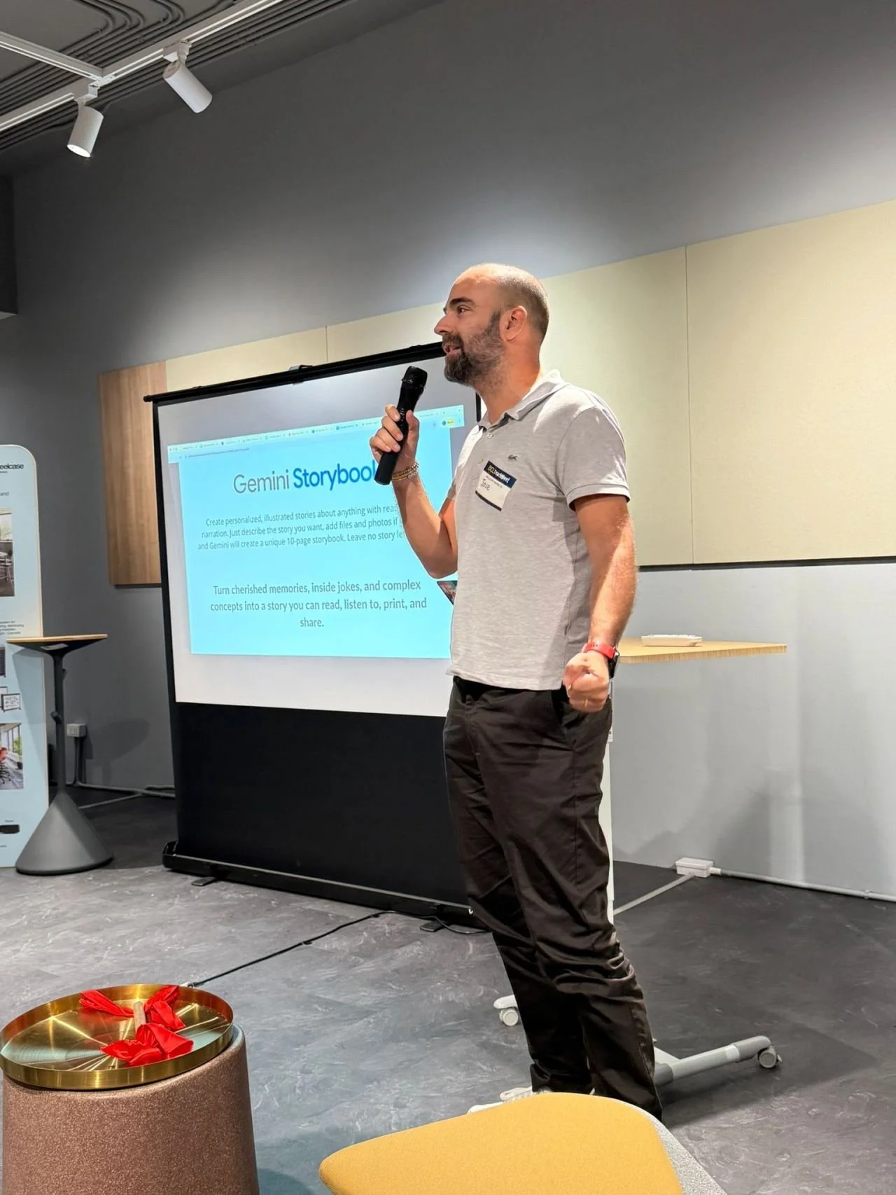 A man holding a microphone and speaking at a presentation in a conference room. There is a projector screen behind him with a slide titled ’Gemini Storybook’. The man is dressed casually in a white polo shirt and brown pants.