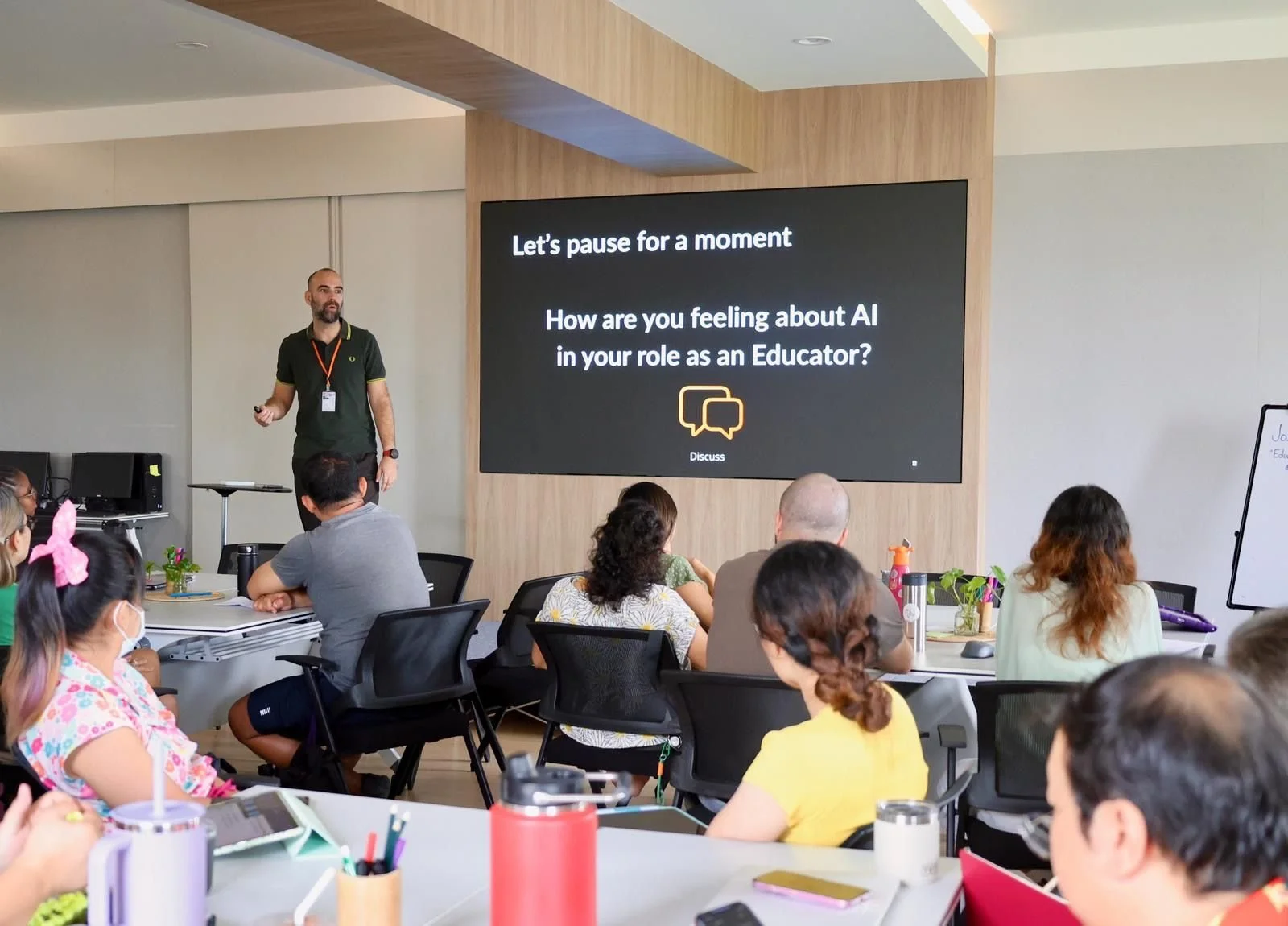 A classroom or conference room with a diverse group of people seated at tables, listening to a presenter standing near a large screen that displays a question about feelings on AI in the role of an educator. The room has computer stations and various items on the tables, with some participants wearing masks.