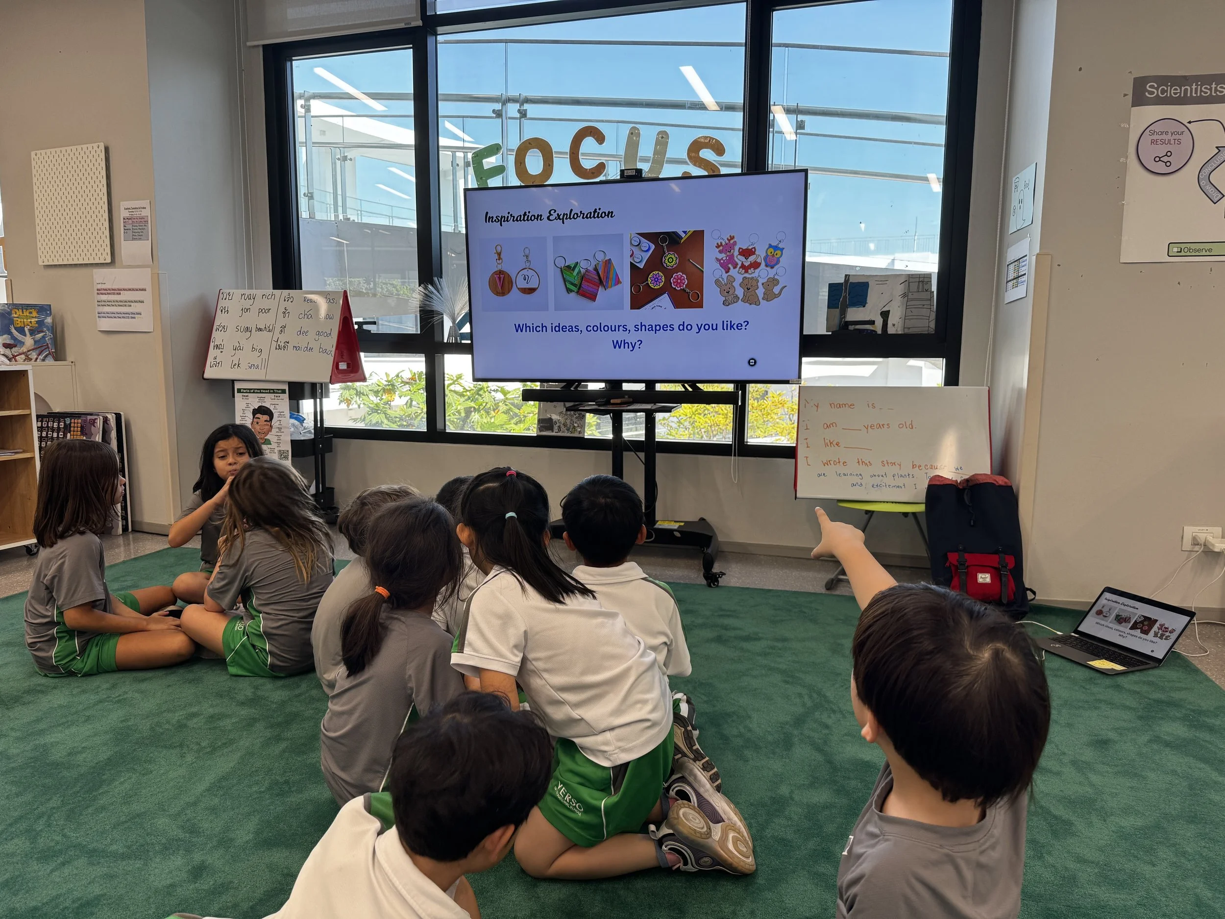 Children sitting on green carpet in a classroom, listening to a presentation via a large screen. The screen displays colorful images and questions about ideas, colors, and shapes.