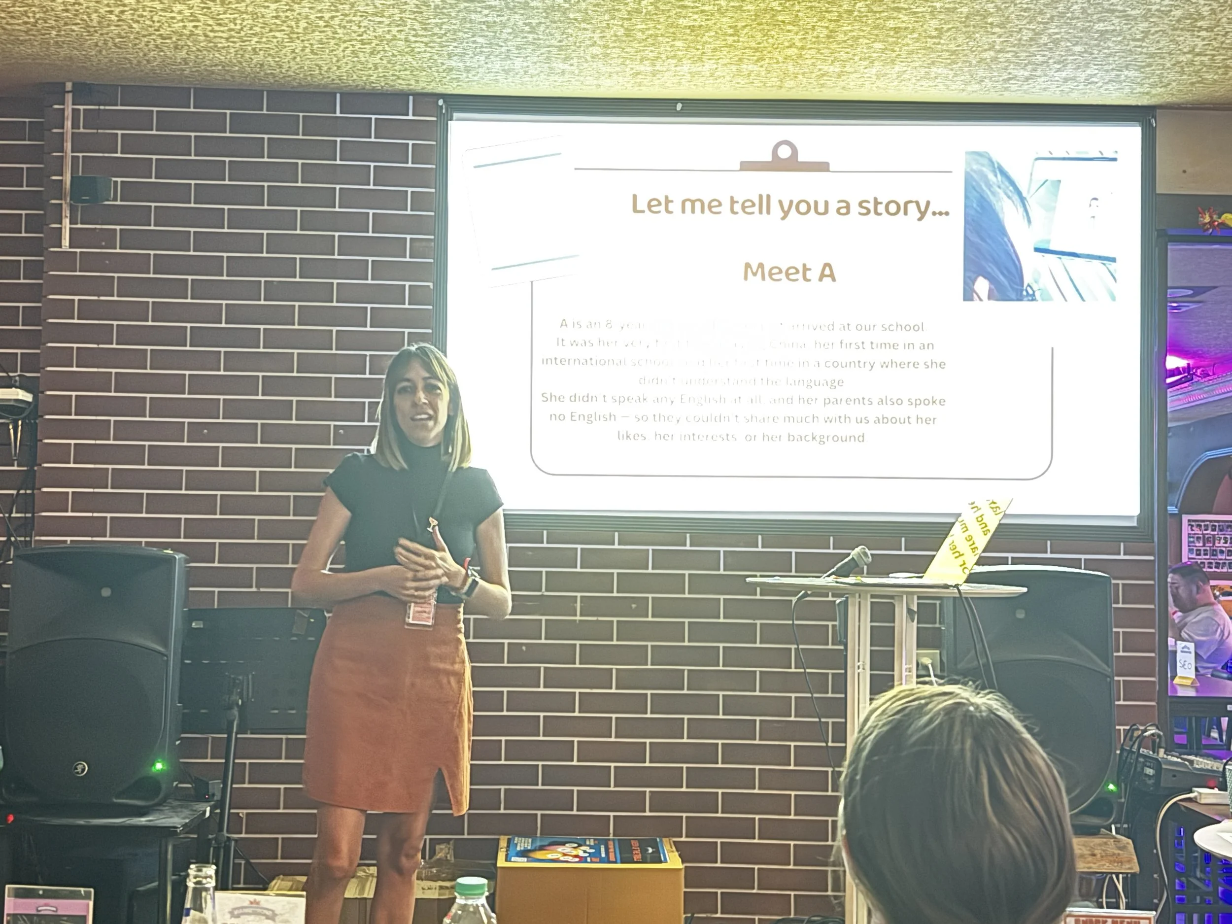 A woman with shoulder-length blonde hair giving a presentation in front of a large screen displaying a story introduction titled 'Let me tell you a story... Meet A'. She is wearing an orange skirt and a black top, standing in a room with a brick wall background and audio equipment around.