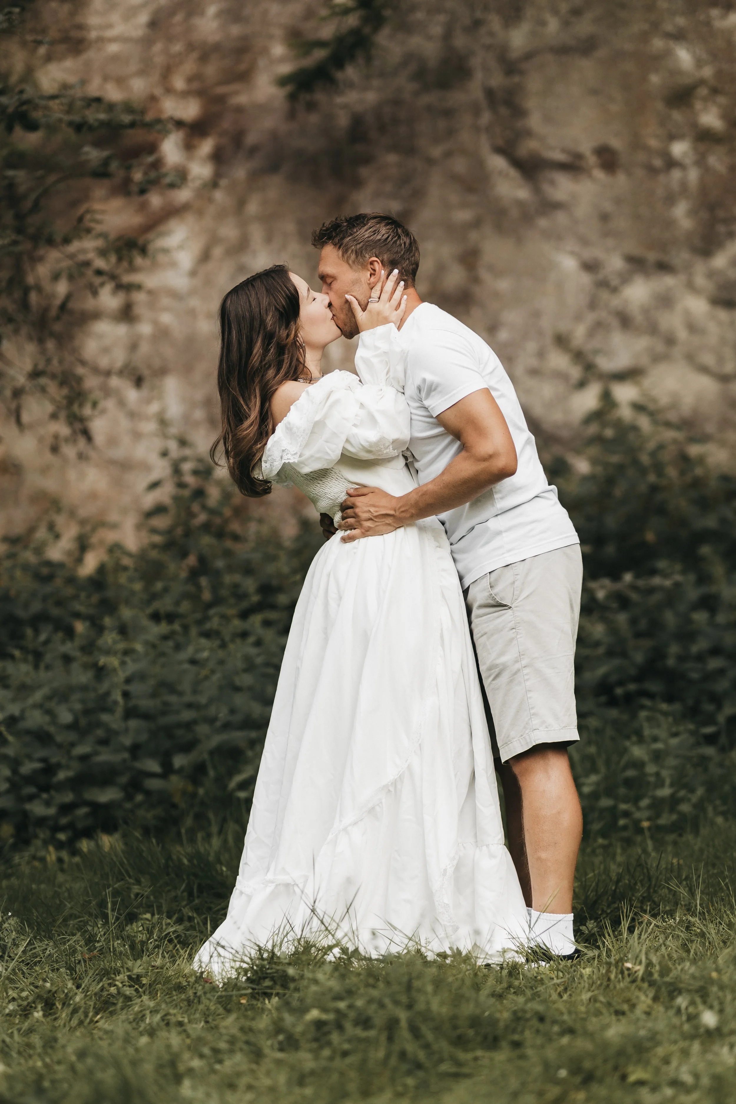 Ein Paar küsst sich in einer natürlichen Umgebung, sie trägt ein weißes Kleid, er trägt ein weißes T-Shirt und Shorts.