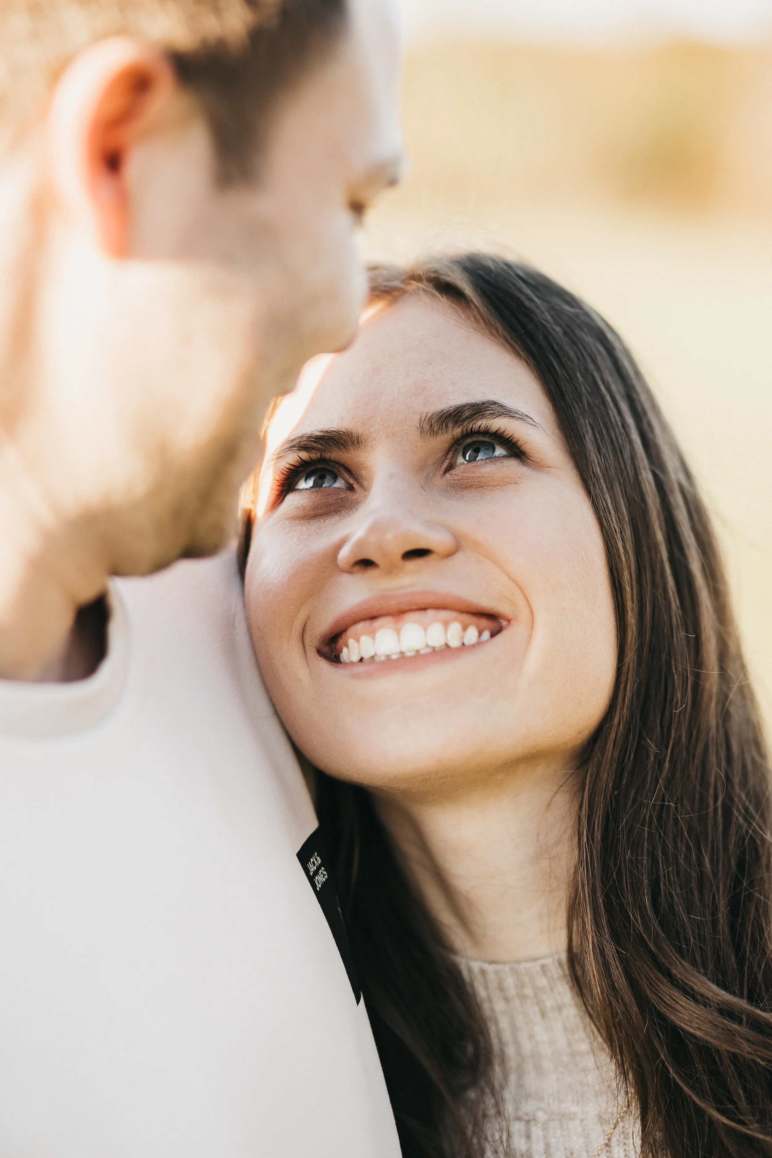 Nahaufnahme eines lachenden Paares, Frau schaut liebevoll zu ihrem Partner, im Hintergrund ein unscharfer heller Himmel.