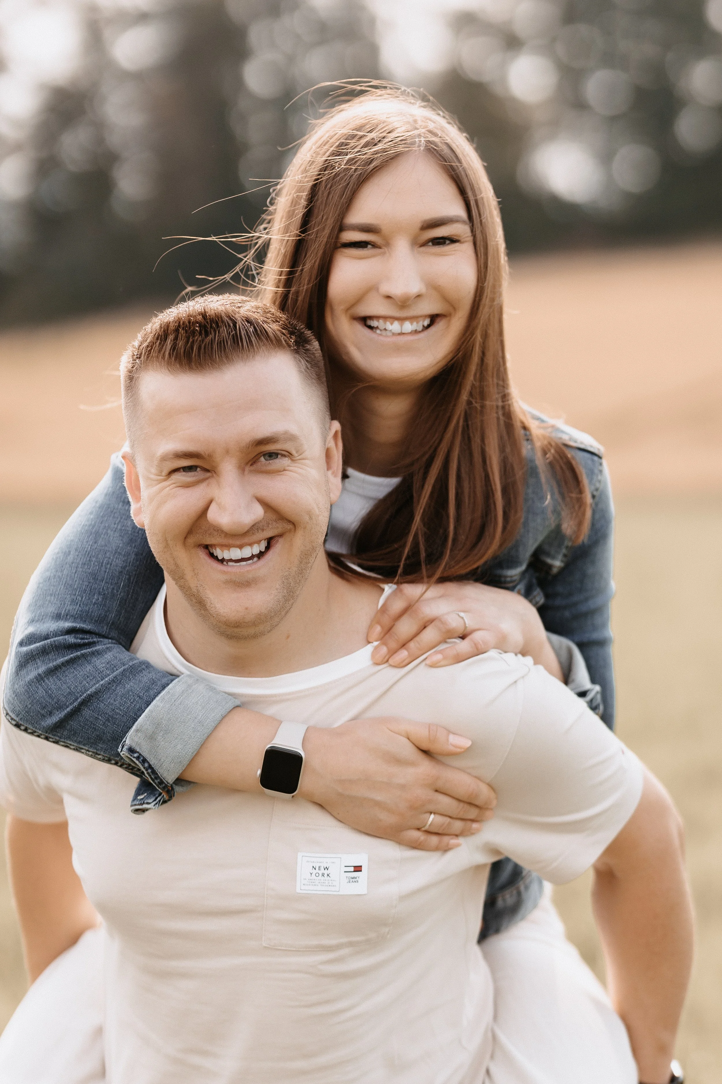 Ein lachendes Paar, die Frau sitzt auf den Schultern des Mannes, im Freien, im Hintergrund Bäume und offener Himmel.