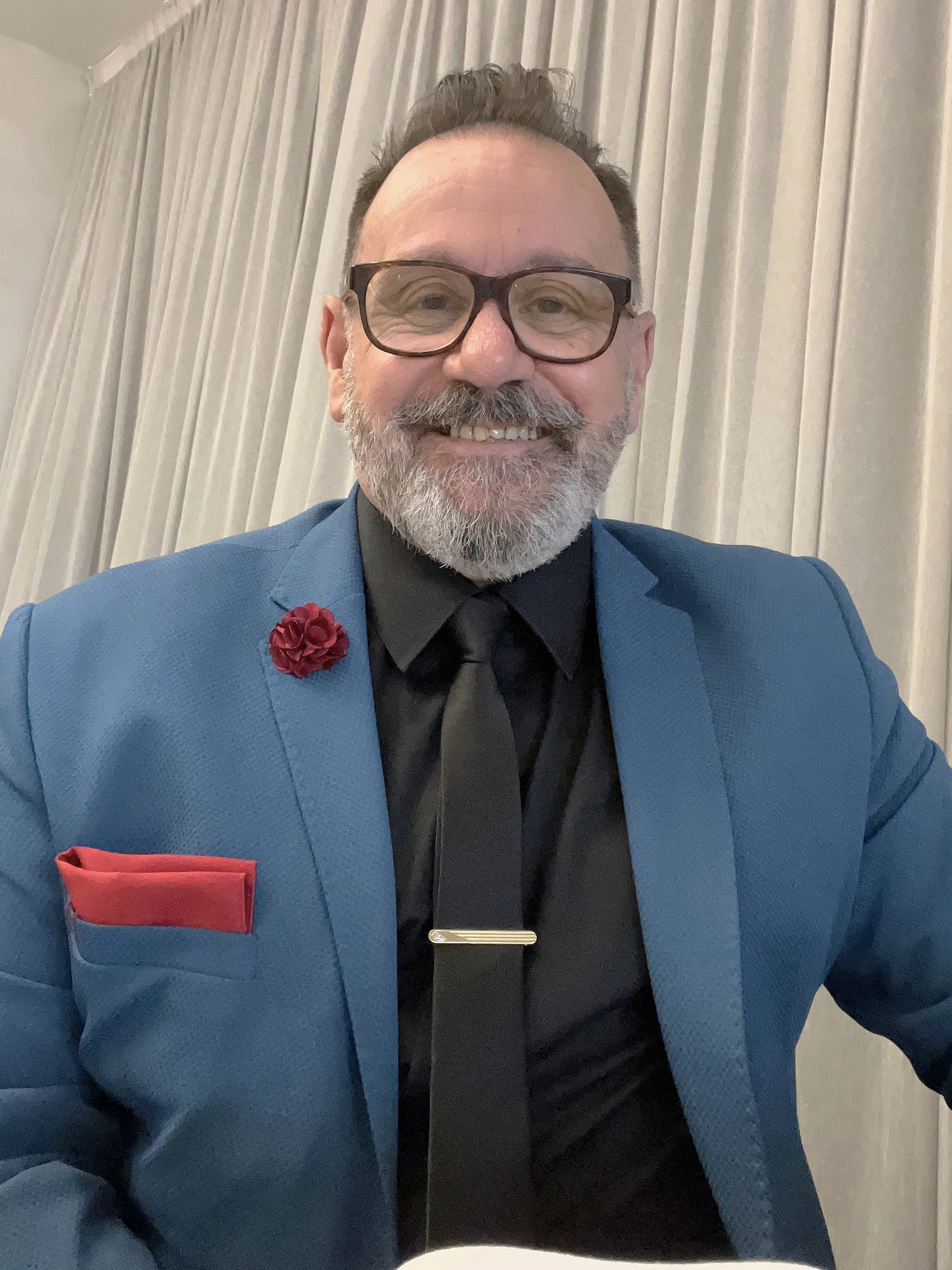 A smiling man with glasses and a gray beard wearing a blue suit, black shirt, and black tie, sitting in front of cream-colored curtains.