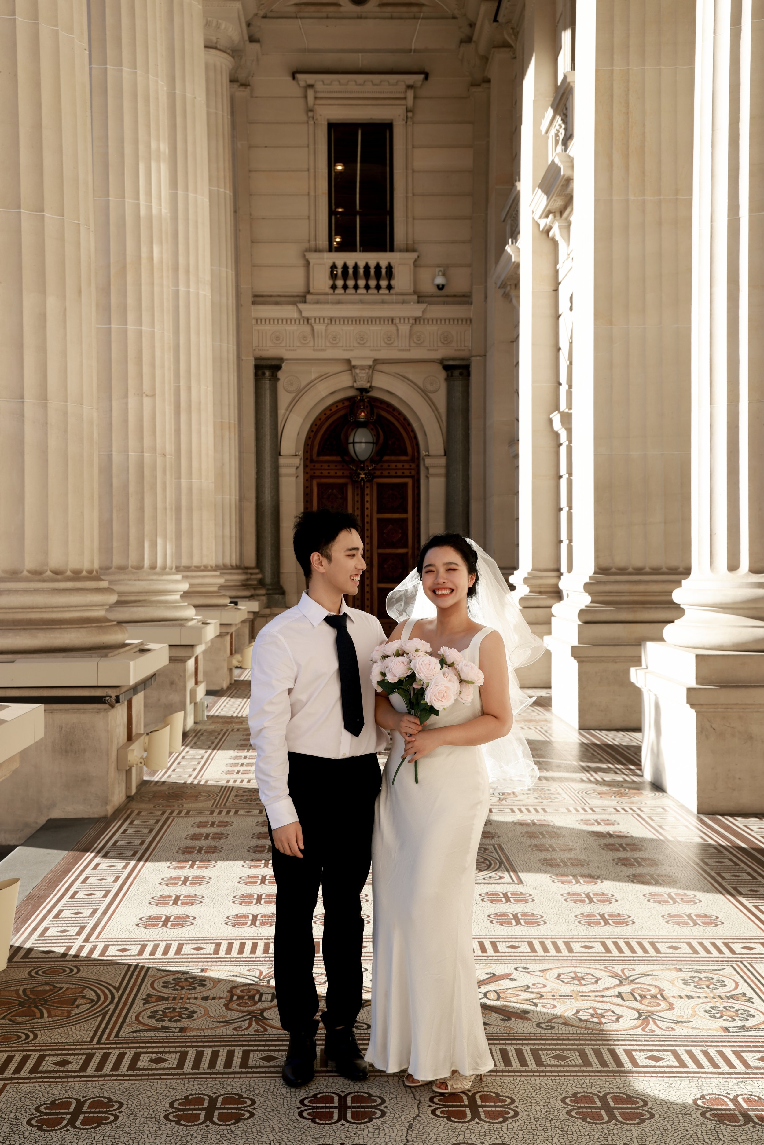 Couple in wedding attire standing inside grand, classical architectural building with large columns and ornate floors, smiling at each other; the woman is holding a bouquet of pink roses, and the man is wearing a white shirt with black tie.