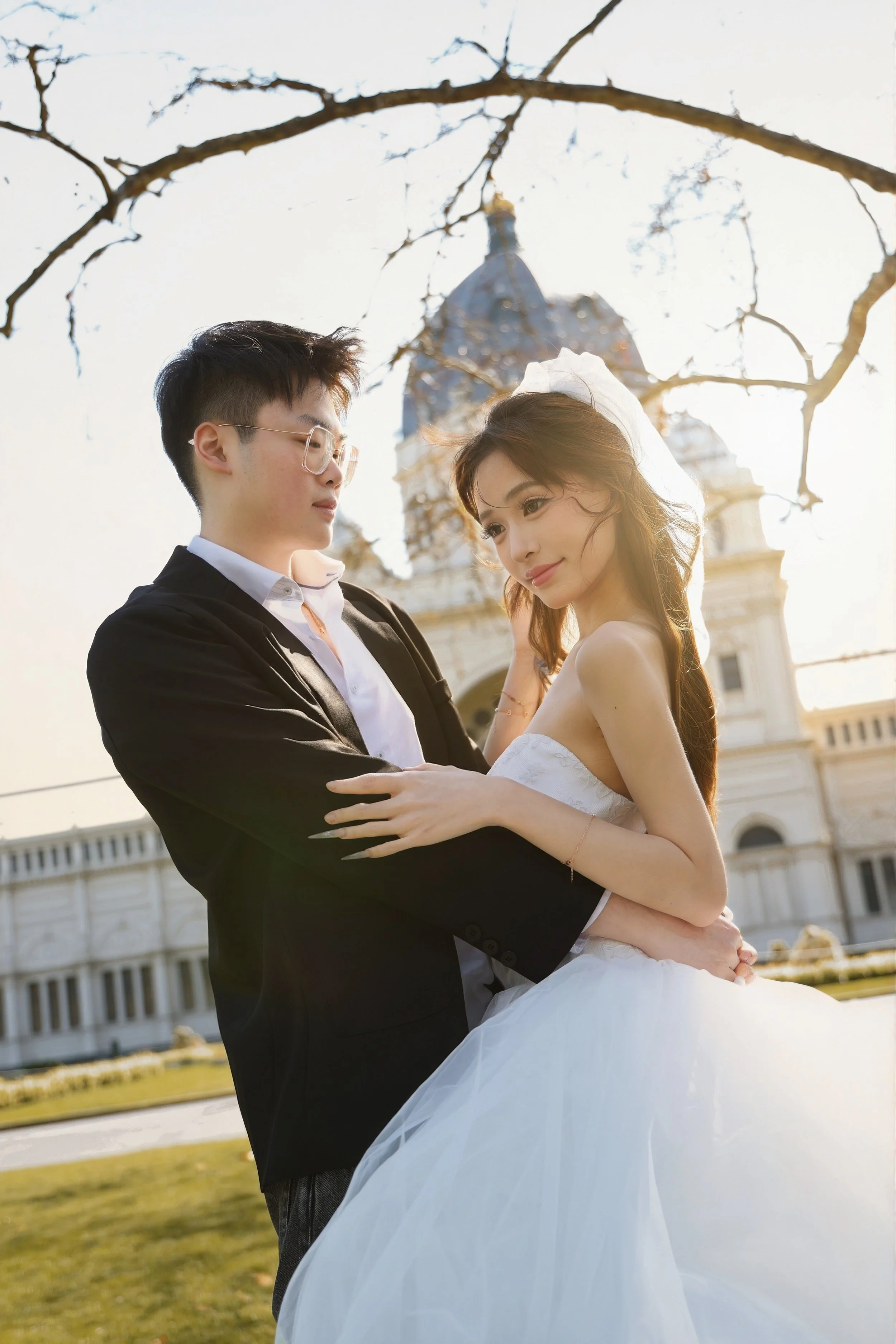 A young couple, dressed in wedding attire, standing outdoors in front of a historic building with a dome, holding each other and gazing at each other during sunset.