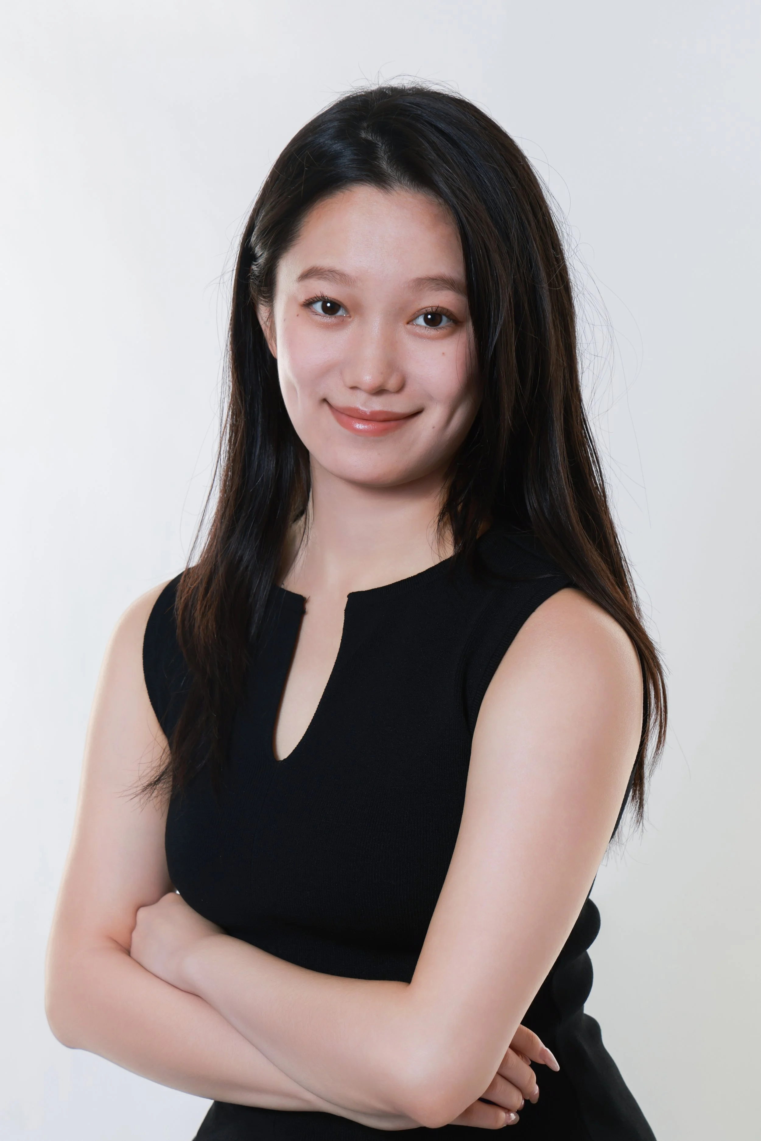 A young woman with long black hair and fair skin, wearing a sleeveless black top, smiling with arms crossed in front of a plain white background.