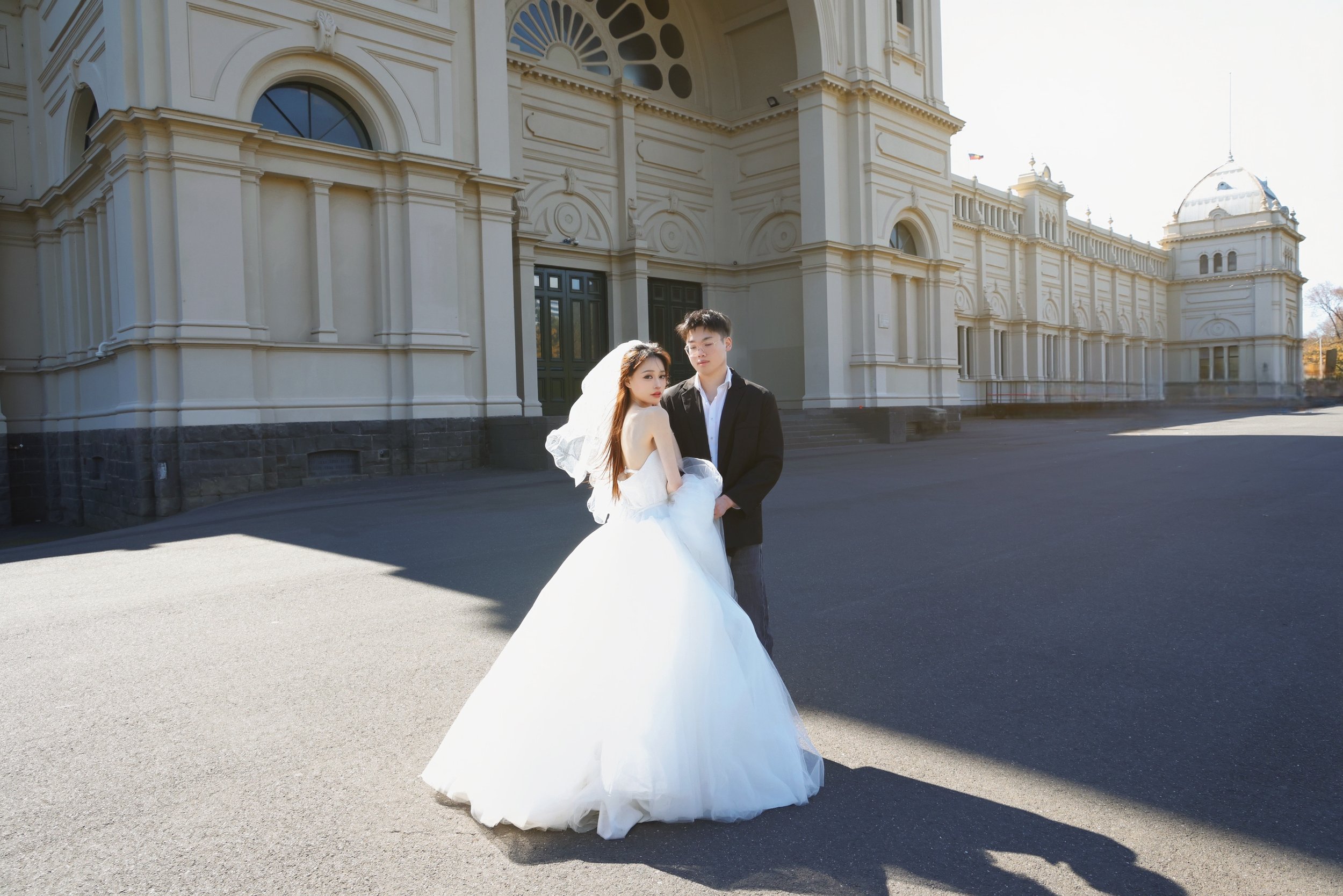 A couple in wedding attire standing outside a large, ornate building with classical architecture. The woman is wearing a white wedding gown and veil, and the man is dressed in a black suit with a white shirt.