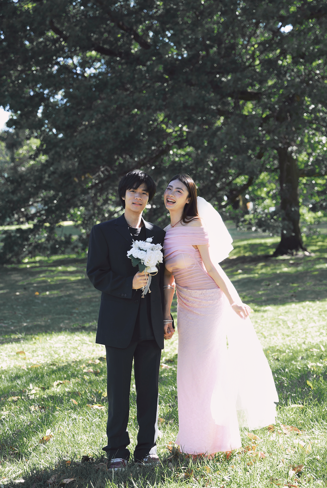 A bride and groom standing outside in a park on their wedding day; the bride is in a pink dress with a veil, and the groom is in a black suit holding a bouquet of white flowers.