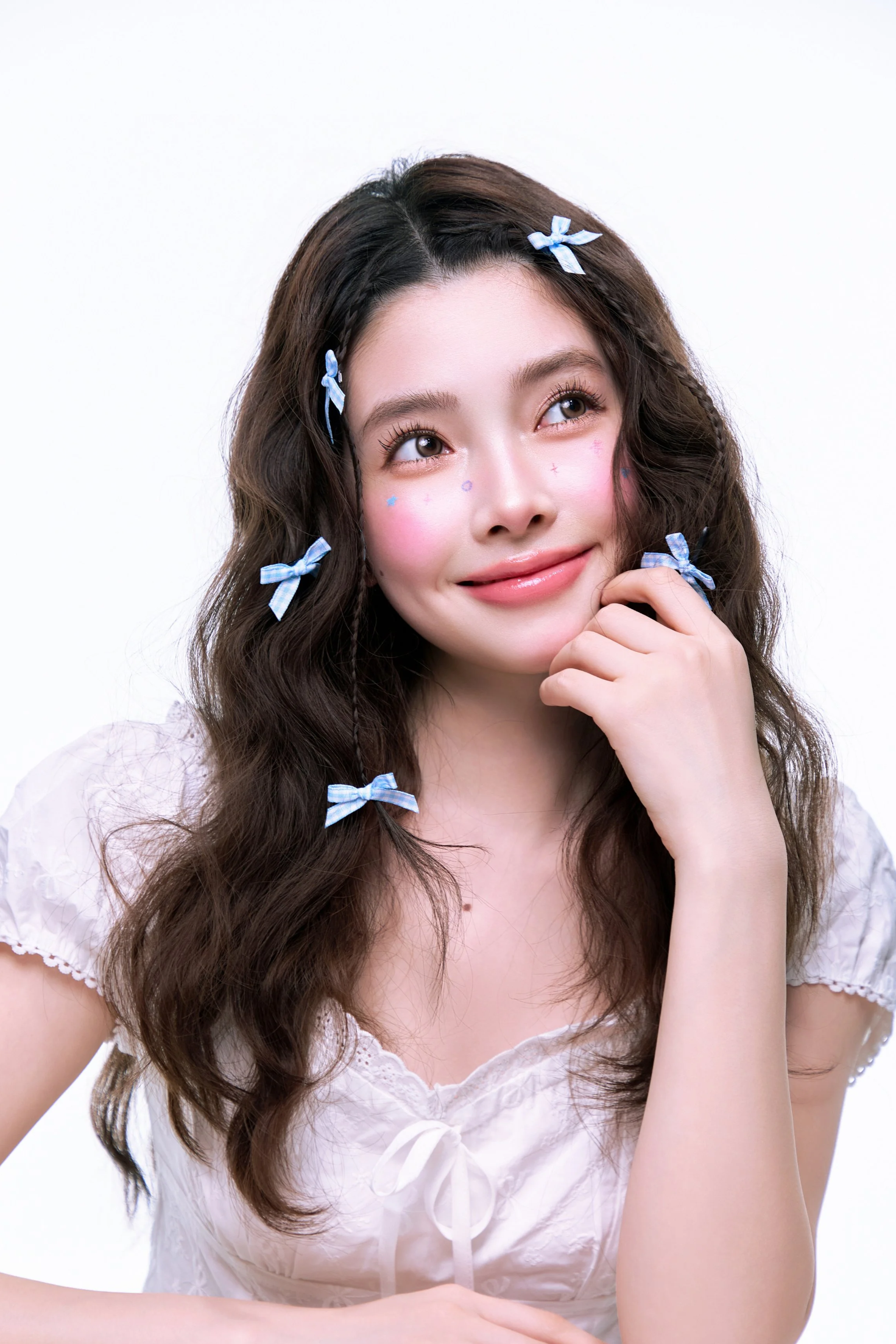 Young woman with long, wavy brown hair decorated with multiple light blue bows, wearing a white blouse with lace details and tie front, smiling gently, with face painted with small, colorful star and heart stickers, posing against a plain white background.