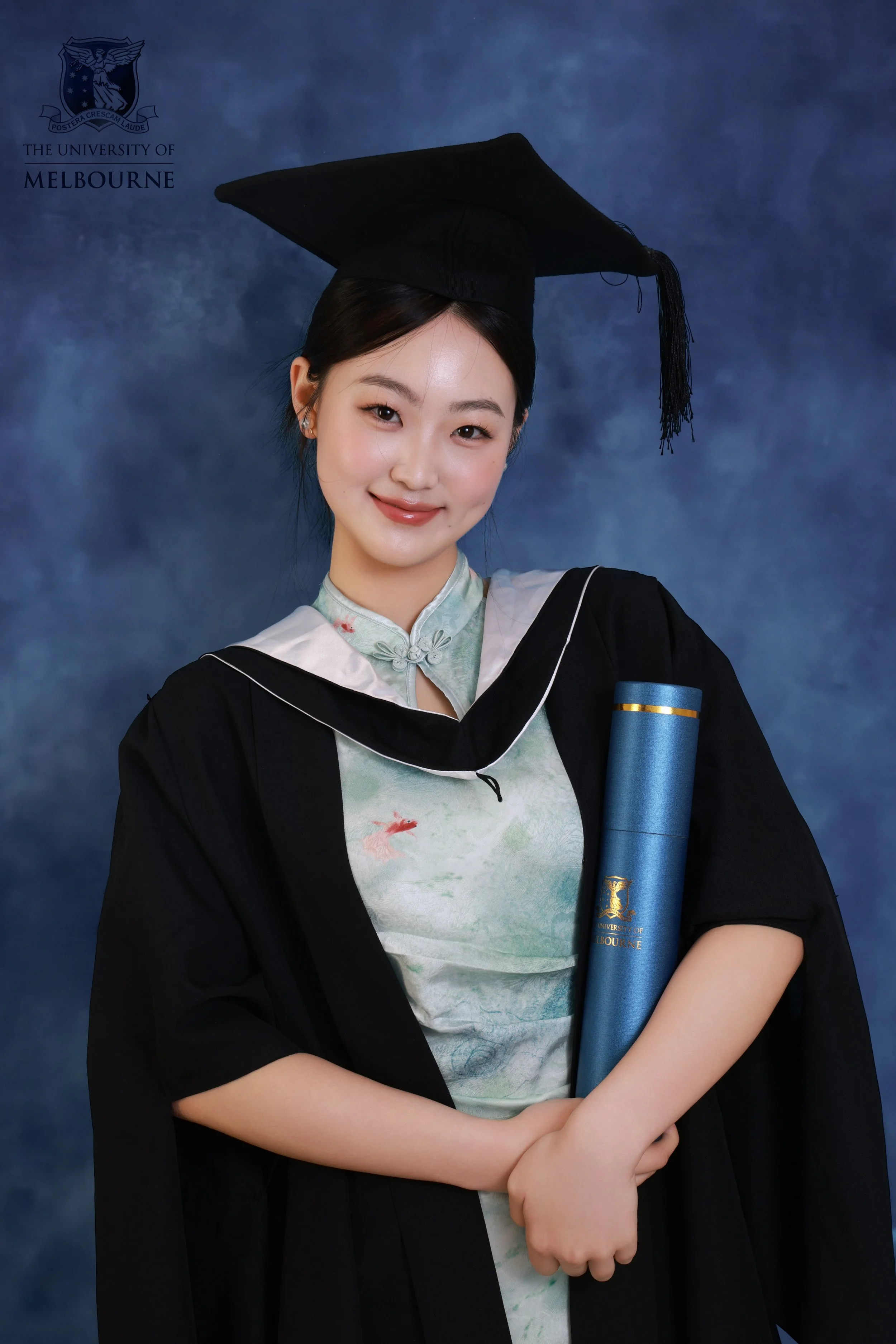 A young woman in graduation attire, wearing a black cap and gown, holding a diploma, and smiling at the camera. The background features the University of Melbourne logo.