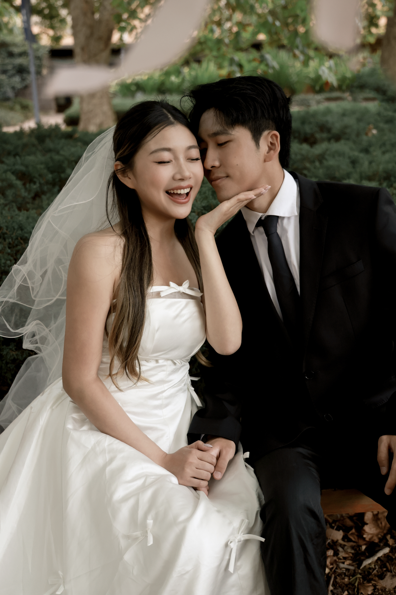A bride and groom sitting outdoors, close together, touching foreheads and smiling. The bride is wearing a white wedding dress and veil, while the groom is in a black suit with a tie. They appear happy and intimate.