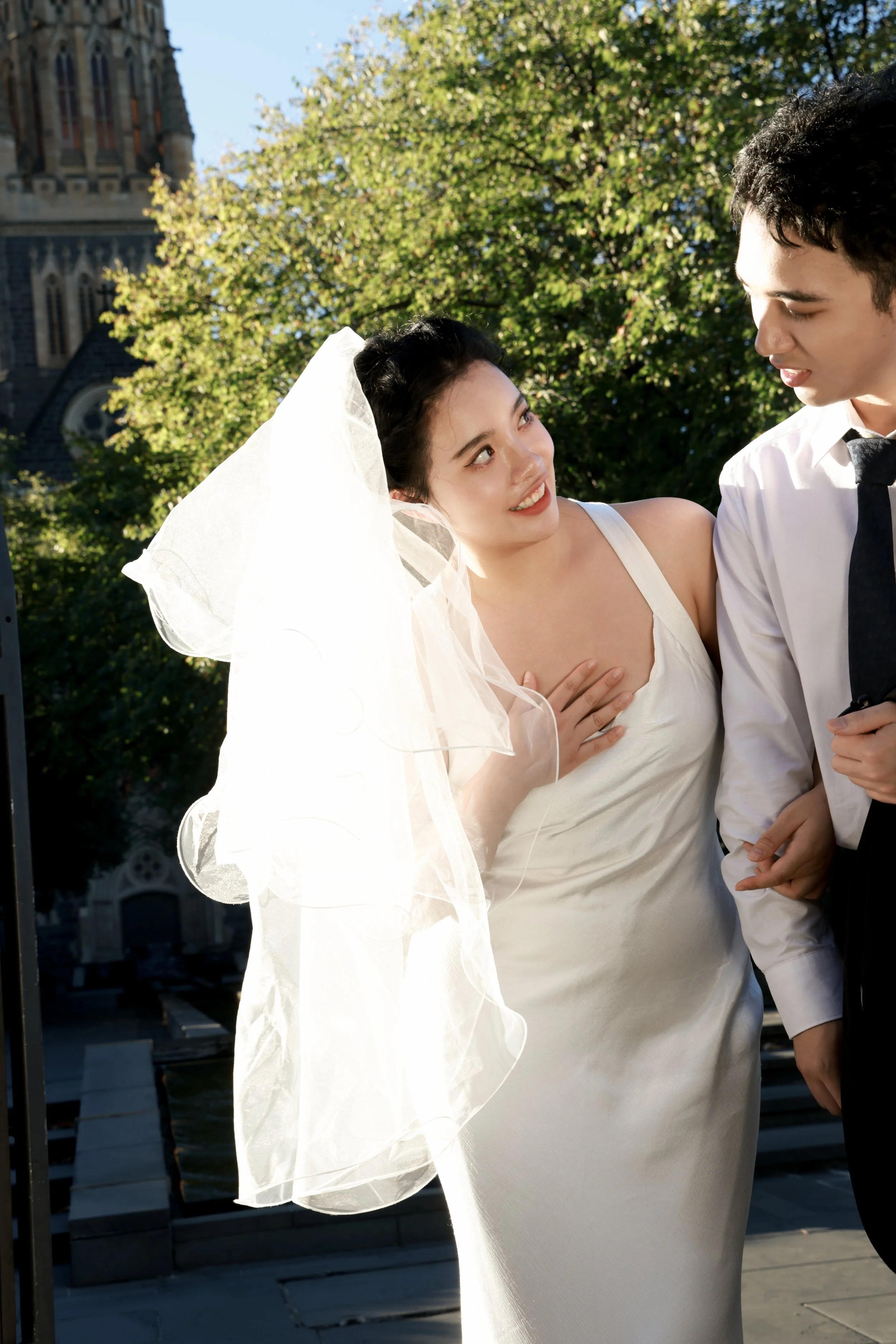 A bride and groom sharing a moment outdoors, with the bride wearing a wedding dress and veil, and the groom in a white shirt and dark tie, standing in front of a church with green trees in the background.