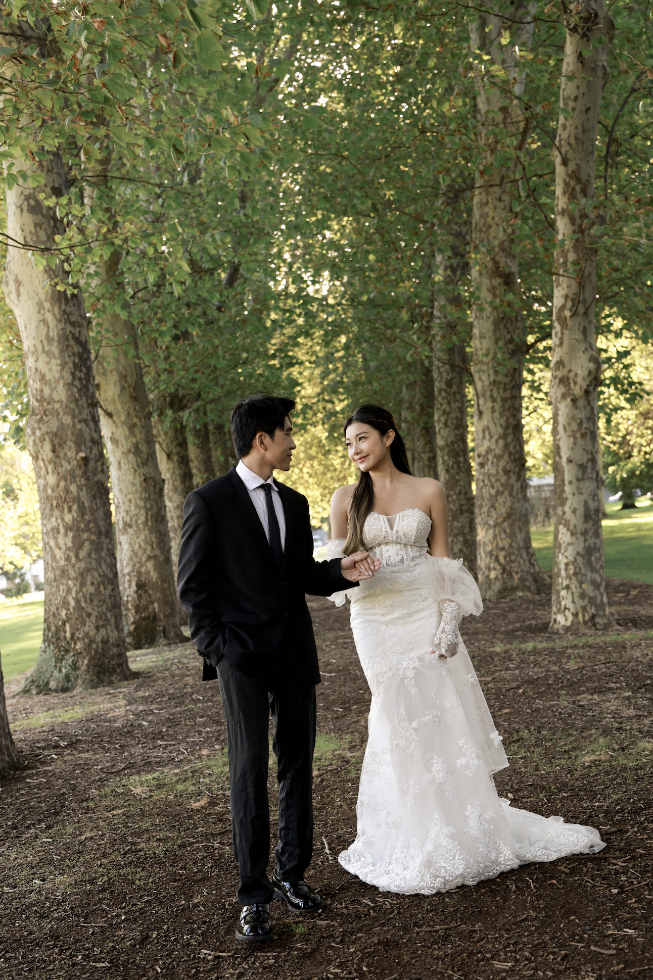 A bride and groom happily walk together under tall trees in a park, with sunlight filtering through the leaves.