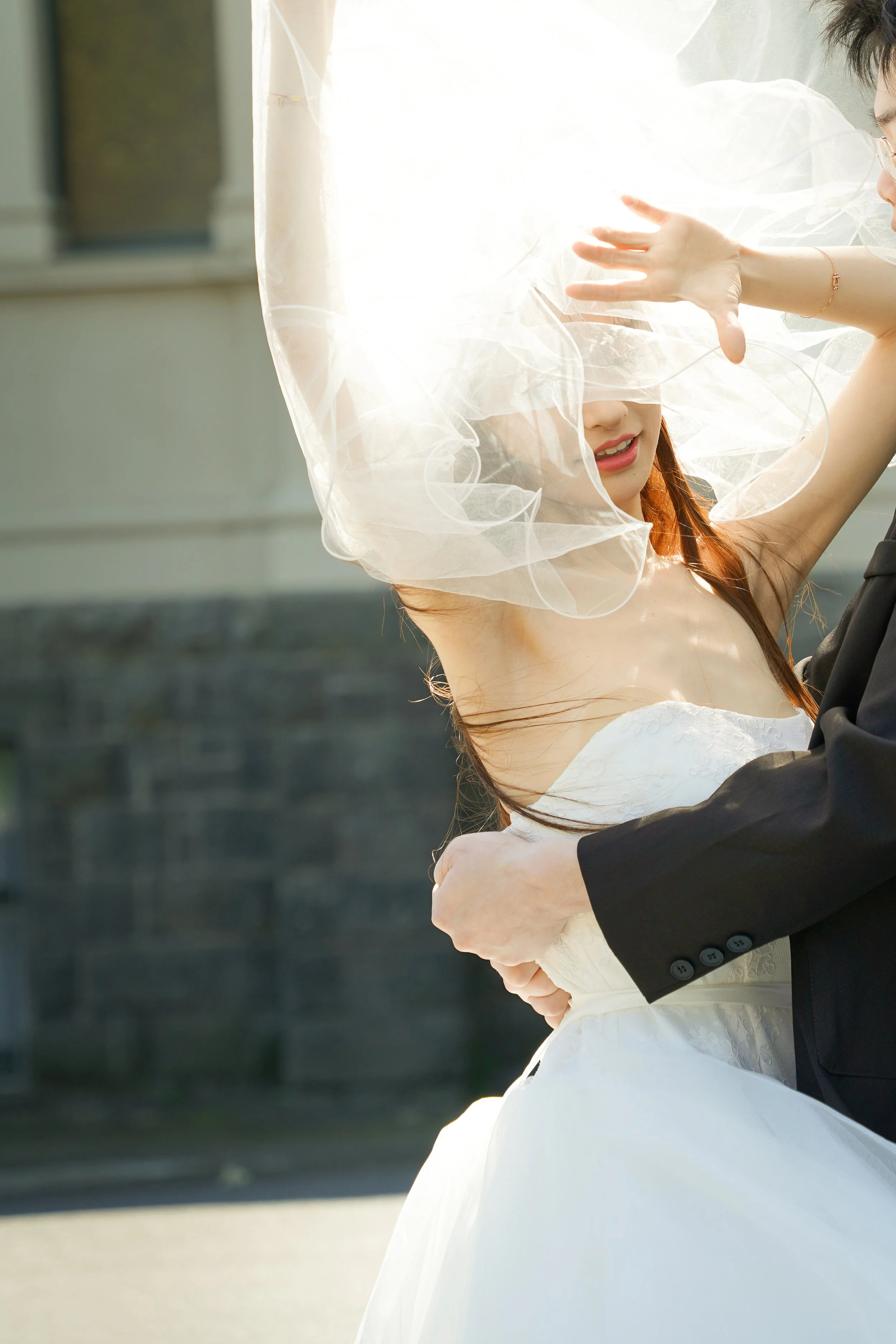 A bride with brown hair in a white wedding dress and veil being embraced and dancing with her groom in a black suit outdoors during daytime.