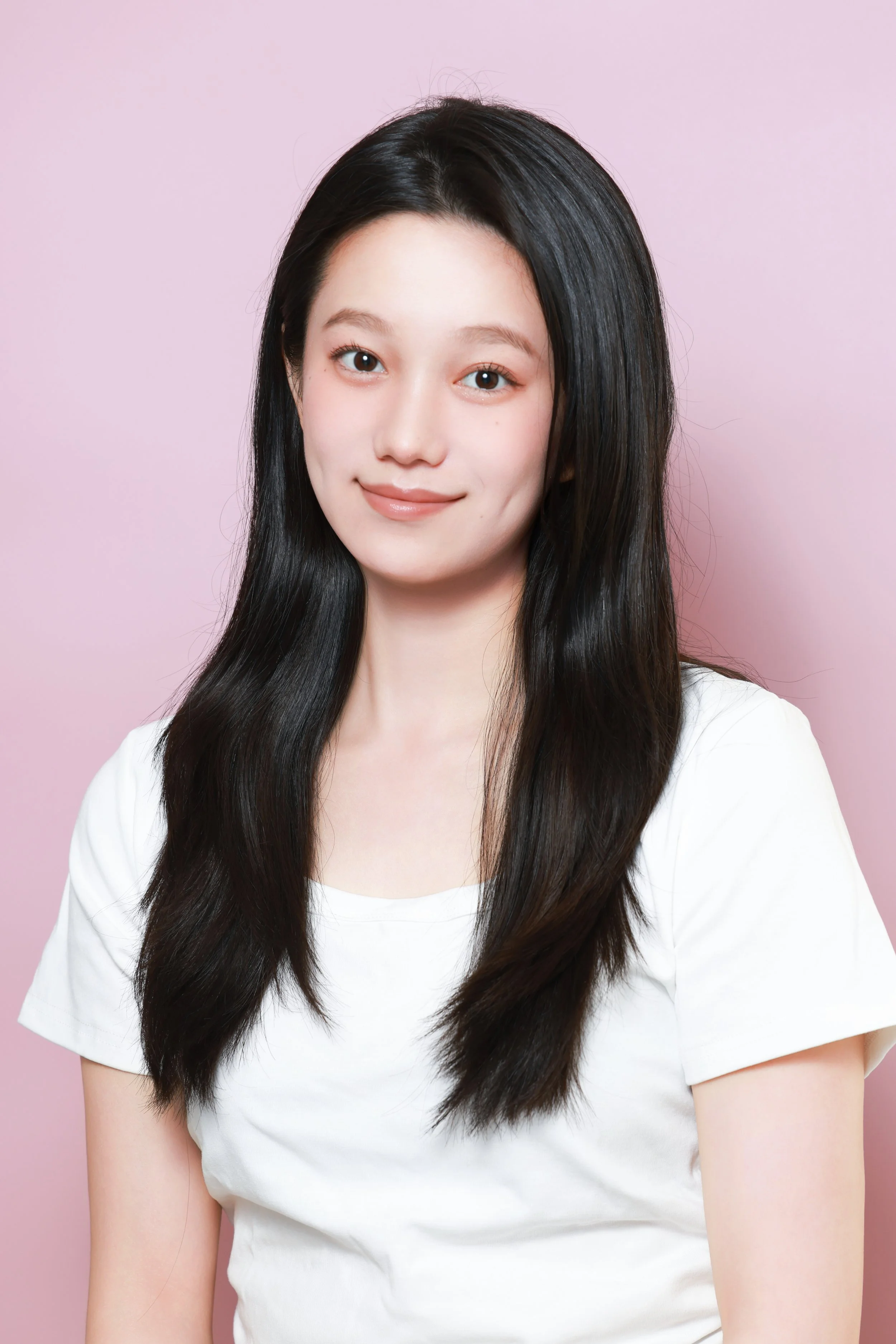 Portrait of a young woman with long dark hair, wearing a white shirt, smiling softly, against a pink background.