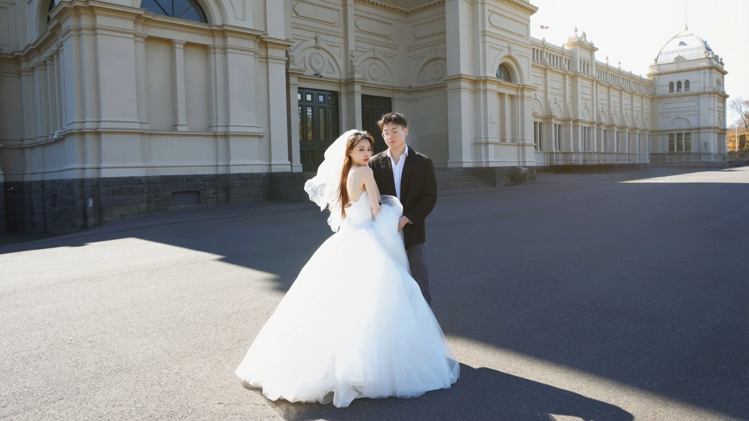 A bride in a white wedding gown with a veil and a groom in a white shirt and black jacket standing together outside on a paved area in front of a large, classical-style building with beige walls and dark green doors, during daytime.